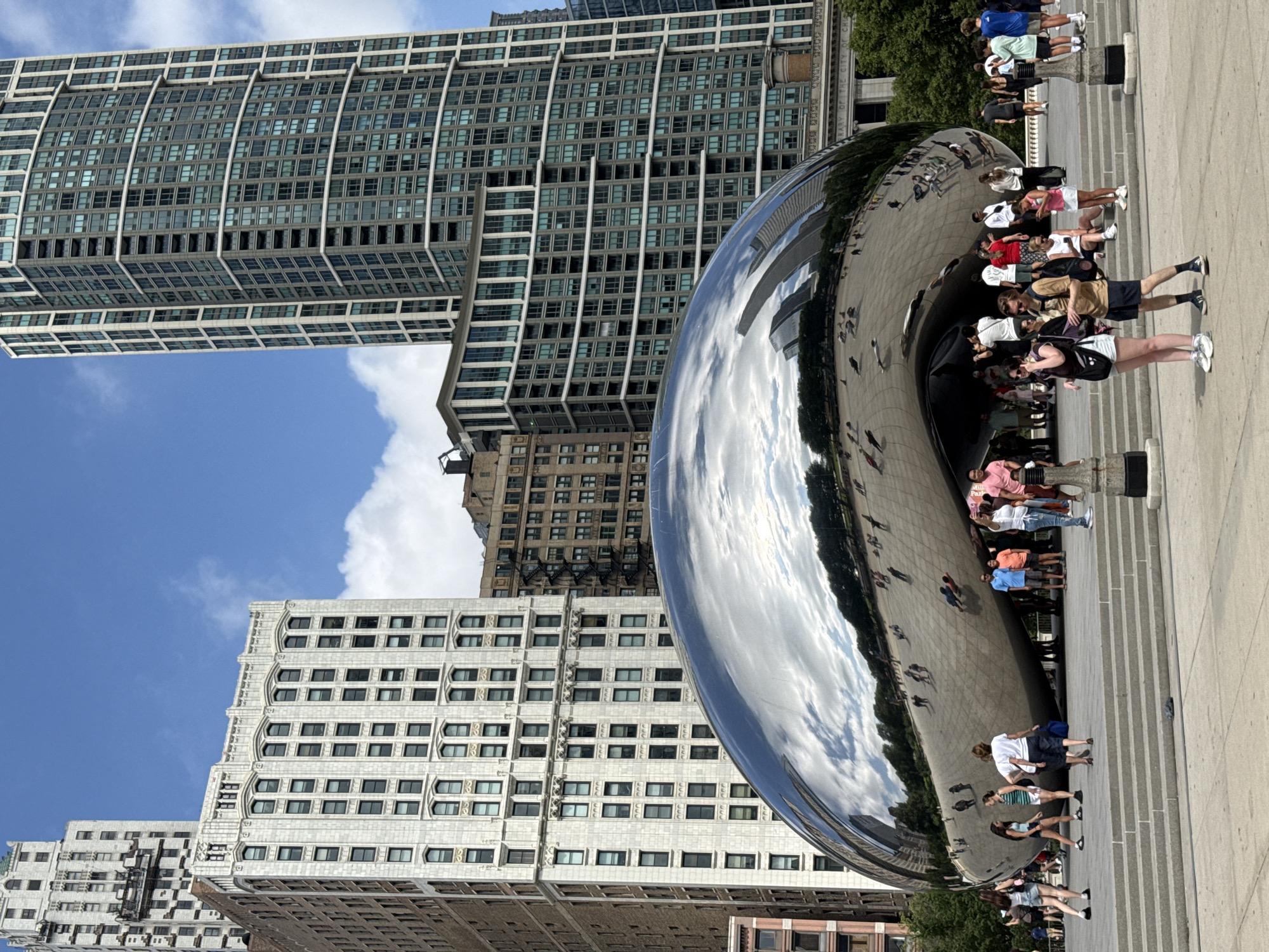This media file captures a daytime scene in Chicago, USA, likely during pleasant weather with a clear blue sky and scattered clouds, as evidenced by the bright lighting and reflections. The primary subject is a large, highly polished, metallic sculpture, reflecting the surrounding urban environment and the sky. The sculpture is abstract and amorphous, with a smooth, curved surface that distorts and duplicates the reflections of buildings and people.
The setting is an outdoor public space, paved with concrete or stone, featuring a wide set of stairs leading up towards the sculpture. Several people are scattered around the sculpture, some posing for photos, others observing the reflections, and some simply walking by. Their attire suggests a casual, summery atmosphere. The reflections in the sculpture reveal towering modern and older architectural structures, characteristic of a major city. Specifically, the reflections show tall buildings with numerous windows, some appearing to be glass-fronted skyscrapers and others with more traditional brick facades.
One of the prominent buildings in the background is a very tall, modern skyscraper with a grid-like pattern of windows. Another building visible is a lighter-colored, ornate, older building with distinct decorative elements around its windows. The reflection of the sky, with its blue hues and white clouds, is vividly captured on the sculpture's surface, indicating a sunny day. The presence of the iconic sculpture, combined with the distinctive architecture, strongly suggests the location is Millennium Park in Chicago, home to "Cloud Gate," also known as "The Bean." There is no explicit text visible in the image that provides further context. The overall mood is one of urban exploration and appreciation of public art.