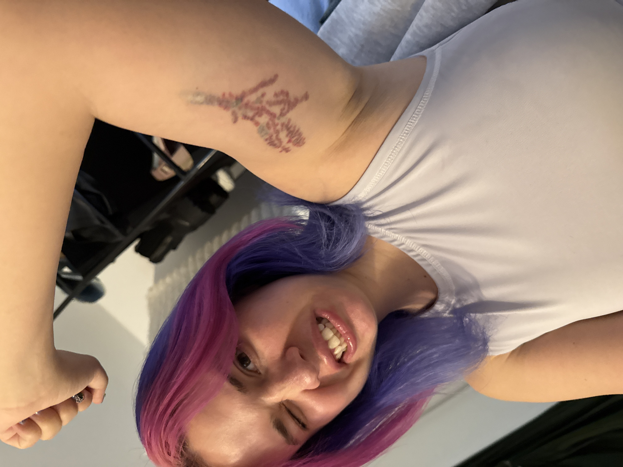 A young woman with striking two-tone hair, featuring vibrant purple and bright pink sections, is captured in a cheerful and confident pose. She faces the viewer with a broad smile, showing her teeth, and winks playfully with one eye. Her right arm is raised and flexed, prominently displaying a delicate, stylized tattoo on her upper bicep.
The tattoo itself depicts a botanical design, possibly a cherry blossom branch or a similar flowering plant, rendered in reddish-purple and muted greenish-grey hues. The intricate details suggest a tree or a complex branching structure with small clusters resembling leaves or blossoms. Faint discoloration or bruising, appearing reddish-purple, is visible around the tattoo, suggesting it may be relatively new or still in the healing process.
The woman is wearing a simple white ribbed tank top, and her left hand is clenched into a fist at the bottom of the frame, contributing to the confident posture. The background is softly blurred and indistinct, indicating an indoor setting. This vibrant portrait captures a moment of self-expression and joy, located in the City of Zagreb, Croatia. No visible text is present in the image.