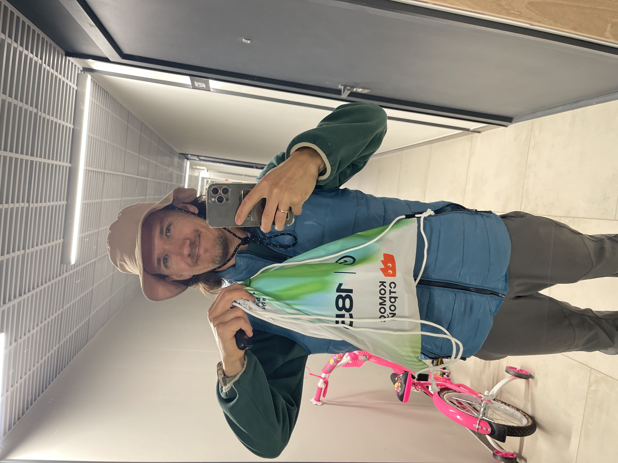 A man is taking a selfie in a hallway, presumably a reflection in a mirror. He is wearing a beige bucket hat, a blue puffy vest over a dark green jacket, and gray pants. He is holding a white drawstring bag with green and white designs and the words "CIBON KOMOD" printed on it. A pink child's bicycle with training wheels is visible behind the bag. The hallway has white tiled walls and a black ceiling fixture with white light panels. The location is Unknown, Unknown.