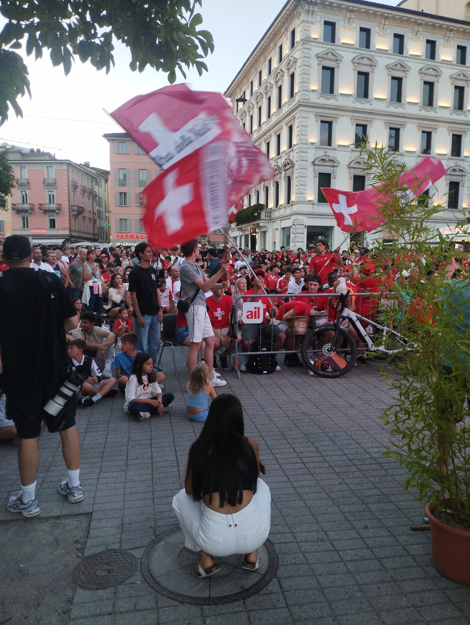 The image depicts a crowd of people gathered outdoors, likely for an event or celebration, as evidenced by the Swiss flags being waved. The architecture in the background, particularly the white building with illuminated windows, suggests an urban setting in a European city. The people are dressed in casual clothing, with many wearing red, a color commonly associated with Swiss national events or sports teams. Some individuals are holding Swiss flags, while others appear to be spectators. A young woman in white pants is crouching in the foreground, facing away from the camera. A potted bamboo plant is visible on the right side of the frame. The ground is paved with cobblestones, and there are some manhole covers visible. The overall atmosphere appears to be festive and communal. The presence of Swiss flags and the architectural style strongly indicate that the location is Lugano, Switzerland, a city known for its Italian influences and scenic setting. The specific building in the background is likely a significant landmark or administrative building in Lugano's city center.