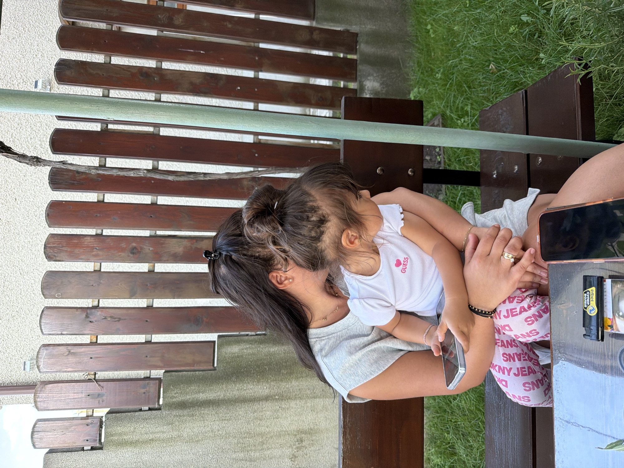 In Burgas, Bulgaria, a young woman with dark hair pulled into a bun, wearing a grey t-shirt, sits on a wooden bench outdoors, embracing a small child. The child, also with dark hair, wears a white t-shirt with the "GUESS" logo and a pink heart. The woman's left arm is around the child, and she holds a silver smartphone in her hand. The child faces away from the camera, looking to the right. Behind them is a wooden fence, and to the left, a textured wall. The ground is covered with green grass, and a table is partially visible in the foreground, with two smartphones and a lighter on it. The scene appears to be a casual outdoor setting, likely a backyard or garden.