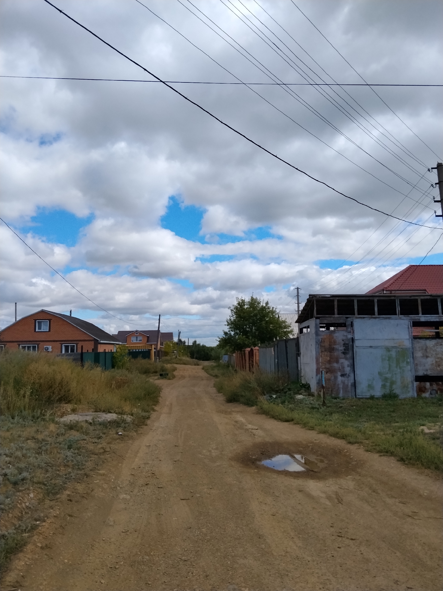 The image depicts a dirt road in a rural or suburban area, likely on the outskirts of Kokshetau, Kazakhstan. The road is flanked by dry grass and vegetation on both sides. On the left, there is a brick house with a dark roof, a white-framed window, and an air conditioning unit. Further down the road, more houses are visible. On the right, there is a partially constructed building with a metal roof and corrugated metal walls, adjacent to a fence. Overhead, multiple power lines crisscross the sky, which is partly cloudy with patches of blue sky visible. A puddle of water reflects the sky on the dirt road in the foreground. The overall impression is of a quiet, undeveloped area.