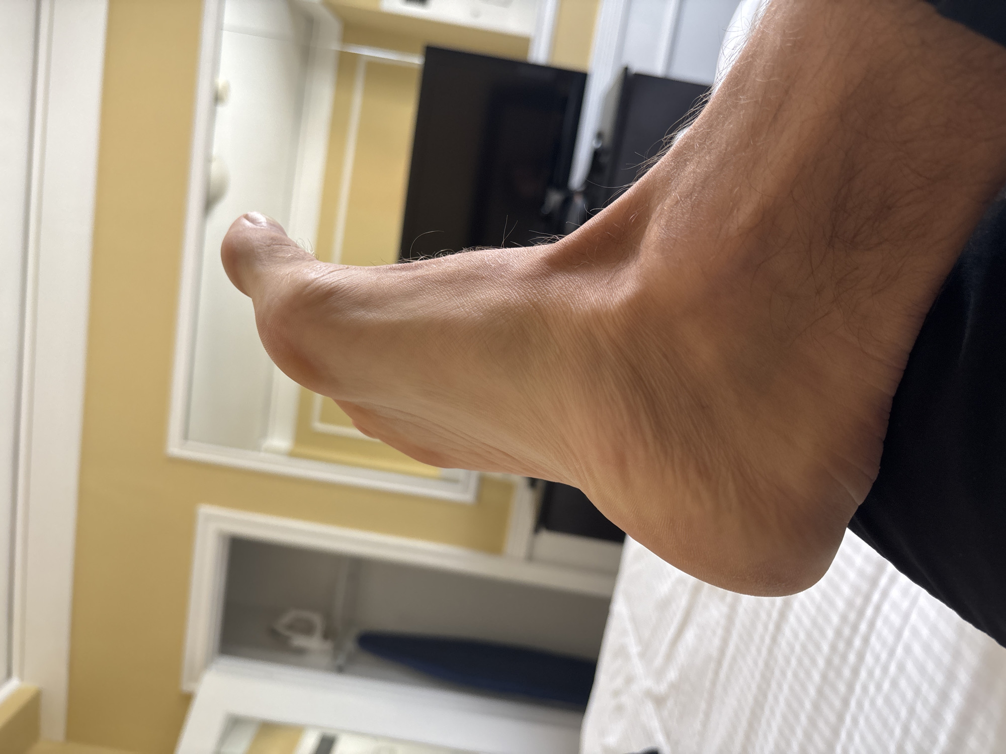 A close-up, low-angle shot captures the side of a bare foot and lower leg, positioned on a black surface that appears to be clothing. The foot is tanned with visible skin texture and fine hairs on the lower leg. The foot is angled upwards, with the toes pointing towards the top of the frame. In the background, a white quilted bed is visible, leading to a yellow wall. A portion of a doorway with glass panes is on the left, revealing an ironing board inside a closet. To the right, a television is mounted on a dark cabinet. The overall setting suggests an indoor environment, possibly a hotel room, but the location in San Francisco, United States of America, cannot be determined from the image alone.