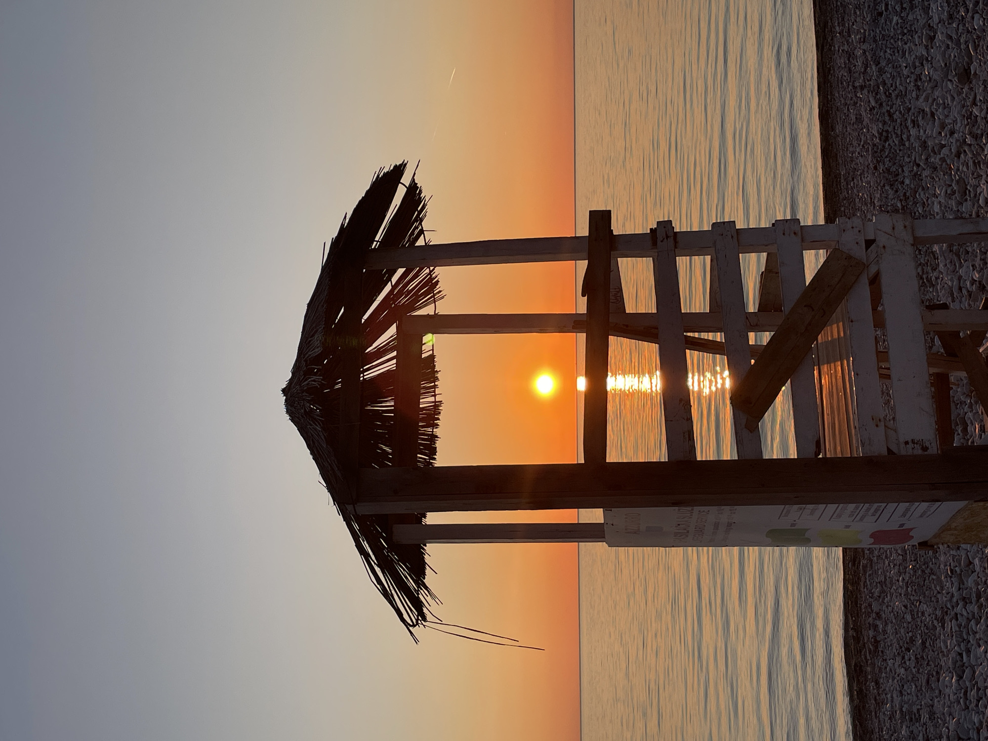 The image shows a lifeguard tower with a thatched roof silhouetted against a sunset over the sea in Bar, Montenegro. The structure is made of weathered white painted wood. The sun is visible between the slats of the tower. The beach is pebbled.