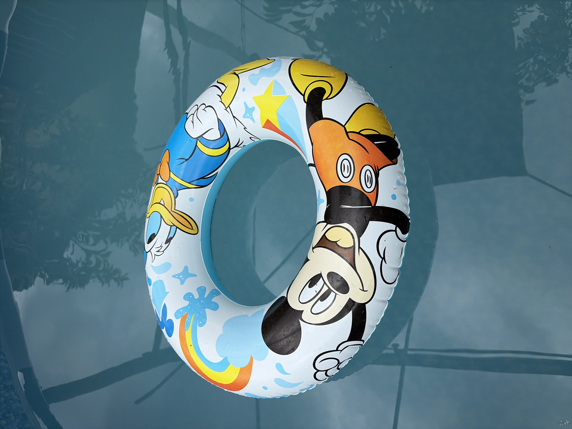 A Mickey Mouse themed inflatable swim ring floats on the surface of a swimming pool in Oxentea, Moldova. The swim ring is primarily white with blue accents and features cartoon characters, including Mickey Mouse and Donald Duck, as well as a rainbow and stars. The water is a clear blue with reflections of overhead foliage and possibly pool structures. The lighting suggests it is daytime.