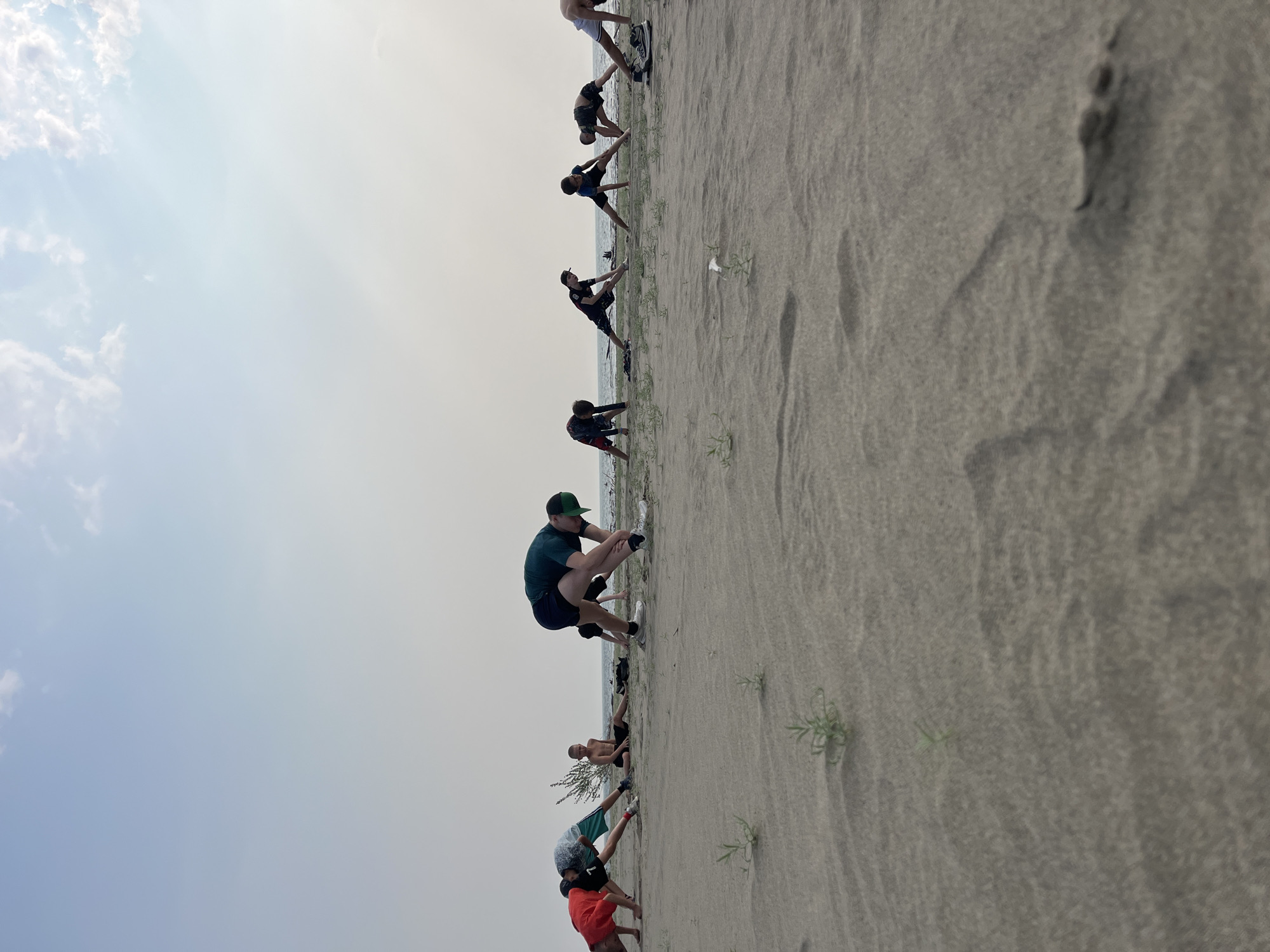 This image depicts a group of people, likely young men and boys, stretching on a sandy beach. They are positioned in a line facing the water, with their bodies bent in various stretching poses. The environment appears to be a natural outdoor setting with sand dunes, sparse vegetation, and a body of water in the background, possibly a lake or river. The sky is overcast with a pale, hazy light. The focus is on the activity of stretching, with the individuals engaged in individual exercises. There are no discernible objects or interactions between the people beyond their shared activity. The scene is devoid of any elements that would suggest a specific urban location or the city of Krasnoturansk, Russia.