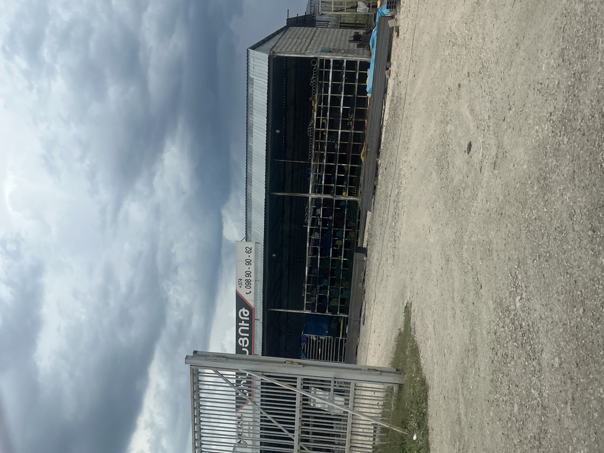 The image depicts an outdoor industrial or commercial storage facility in Chiva, Armenia, under a heavily overcast sky. A single-story building with a corrugated metal roof and concrete block walls dominates the midground. Its front is open, revealing multiple metal racks filled with various metal profiles, pipes, and construction materials. Blue and possibly yellow tarpaulins are visible within the storage area and around the building.
A prominent white and red sign is affixed to the left side of the building's facade. It displays an Armenian script logo, appearing as "ՀՅԱՆԴԱ" (likely 'HYANDA'), and provides contact details: "+374 098 90 - 90 - 62". The "+374" confirms the Armenian location.
The foreground and right side of the image consist of a dusty, gravelly ground. A partially open metal gate with vertical bars is visible in the lower left corner, casting a soft shadow. The sky is filled with dark, dense clouds, suggesting an overcast day with diffused lighting. No human activity is discernible; the scene is static, representing a functional materials storage or distribution point. The time of day is daytime, likely mid-day or afternoon given the soft, ambient light.