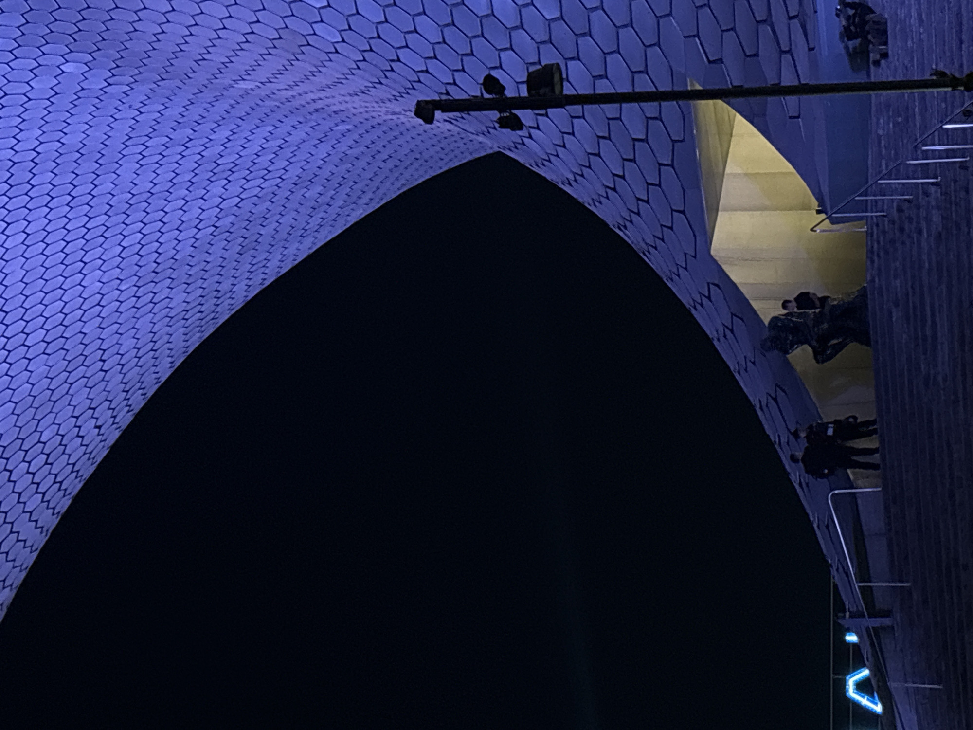 The image shows the Museo Soumaya in Mexico City, Mexico at night. The building features a distinctive, curved exterior covered in hexagonal panels, illuminated with blue light. A metal structure with spotlights is affixed to the exterior. On the right side of the frame, there are sections of a wall with stairs or ledges where several figures can be seen. A blue neon 'V' shape is also visible towards the bottom right. A large dark void fills the interior space of the building's arch, creating a high-contrast composition.