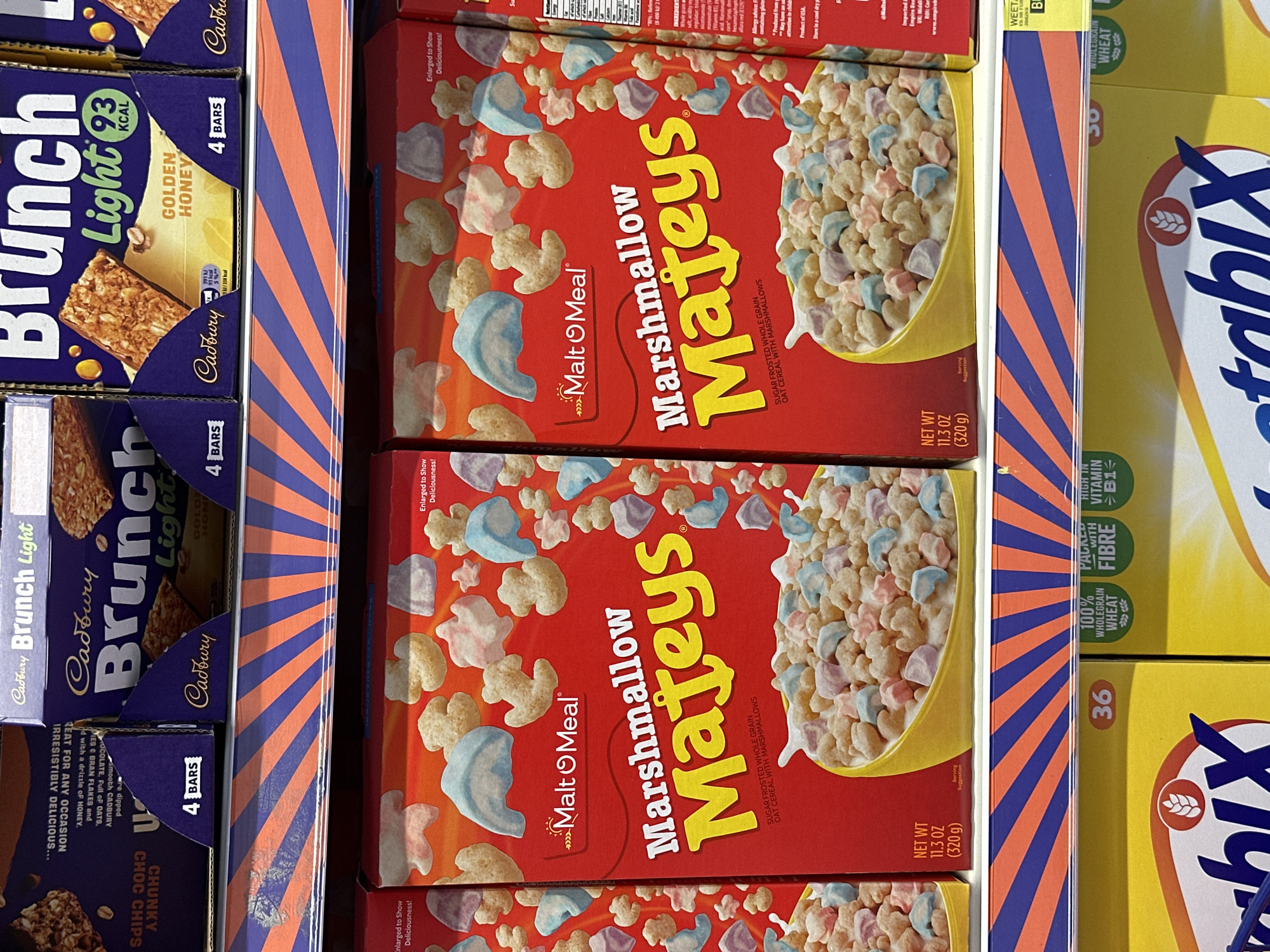 The image presents a close-up of a retail shelf displaying breakfast and snack items. Visible are boxes of "Marshmallow Mateys" cereal, "Cadbury Brunch Light" bars in "Golden Honey" and "Chunky Choc Chips" flavors, and "Weetabix" cereal. The scene likely takes place in a supermarket or grocery store in Wealden, United Kingdom. No people are visible, and the focus is on the products and their packaging. The visual environment is colorful and brightly lit.