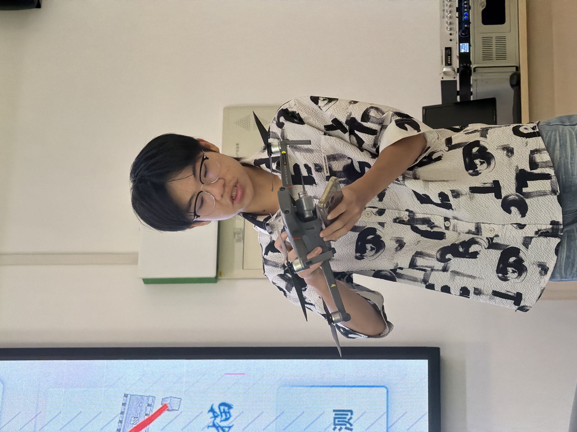 In a classroom or presentation space in Jiangjin, China, a young person wearing a patterned shirt and glasses holds a drone in their hands, appearing to explain or demonstrate its features. The drone, a DJI Mavic 2 Pro, is held horizontally, with its propellers visible. To the left, a large screen displays Chinese characters and a diagram. In the background, shelves hold electronic equipment. The lighting is bright and even.