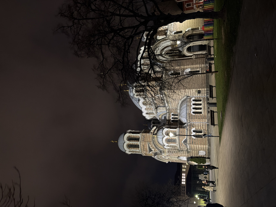 The image depicts a nighttime scene centered on the Church of St Nicholas the Miracle-Maker (Russian Church) in Sofia, Bulgaria. The church, constructed in Neo-Russian style, features a brown and light-striped brick facade, multiple silver-grey domes topped with golden Orthodox crosses, and numerous arched windows, all illuminated by artificial light against a dark, overcast sky.
In the foreground and midground, a dark paved pathway extends from the left. Several individuals are present: a person partially visible on the far left walking right, a group of three individuals walking leftward away from the church, another person walking left, and a lone figure in the mid-left walking away from the camera. Artificial streetlights with white luminaires illuminate the pathways and parts of the church.
To the right of the paved area, a grassy strip contains four dark park benches with slatted seats; one bench on the mid-right appears to have a person seated on it. Bare deciduous trees with intricate branch patterns are scattered, with a large, dark tree dominating the right side of the frame. In the far right, a low, multicolored fence with vertical slats (red, orange, yellow, green, blue, purple) is visible. A building with illuminated windows is discernible in the far left background. The overall illumination is artificial, creating strong contrasts.