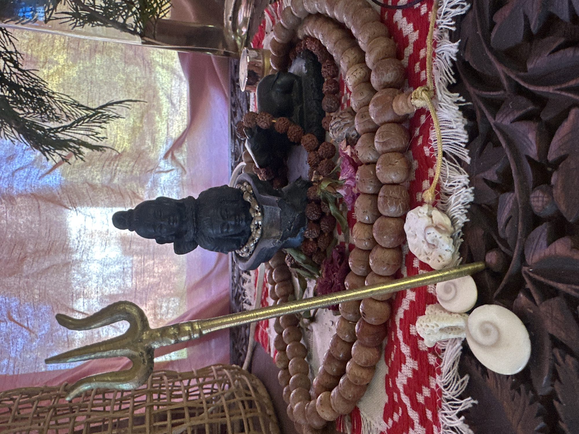 This is a close-up, eye-level, indoor shot of an altar arrangement. The arrangement is set on a dark wooden surface with carved floral motifs, covered partially by a red and white patterned cloth with white fringing. Dominating the foreground are several nautilus-shaped shells and a piece of white coral. A brass trident stands vertically in the center of the frame, its prongs pointing upwards. Behind the trident, a multi-faced black statue, possibly of a deity, is centrally placed. Around the statue, several strands of wooden beads are laid out, some encircling the statue and others fanned out across the cloth. To the right of the statue, a small black figurine of a bull is visible, also adorned with beads. A woven basket is positioned to the left of the trident. In the background, a textured pink fabric hangs loosely, with a sprig of evergreen foliage visible in the upper right corner. The lighting appears to be warm and diffused, suggesting an indoor setting, possibly illuminated by natural light filtering through a window or by artificial lights. The overall arrangement suggests a spiritual or devotional space. The location is not specified in the image, and therefore, cannot be determined to be Thora, Australia.