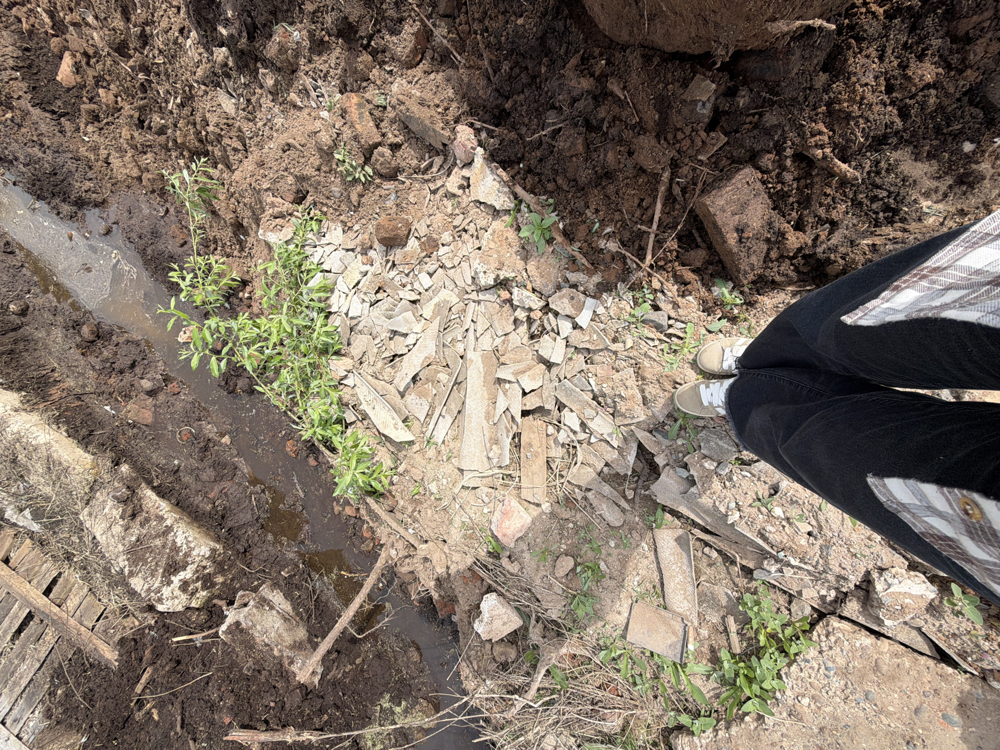 The image shows a patch of disturbed ground, likely in a rural area. The ground is composed of dirt, broken pieces of concrete, and brick fragments. There is a narrow trench with stagnant water running along one side. Several small plants are growing amidst the debris. A person wearing black pants and light-colored sneakers stands near the edge of the disturbed area. The location, Uva, Russia, is outside of the view frame, and the image does not contain any indication that the photograph was taken here. There is no activity or apparent interaction occurring within the frame.