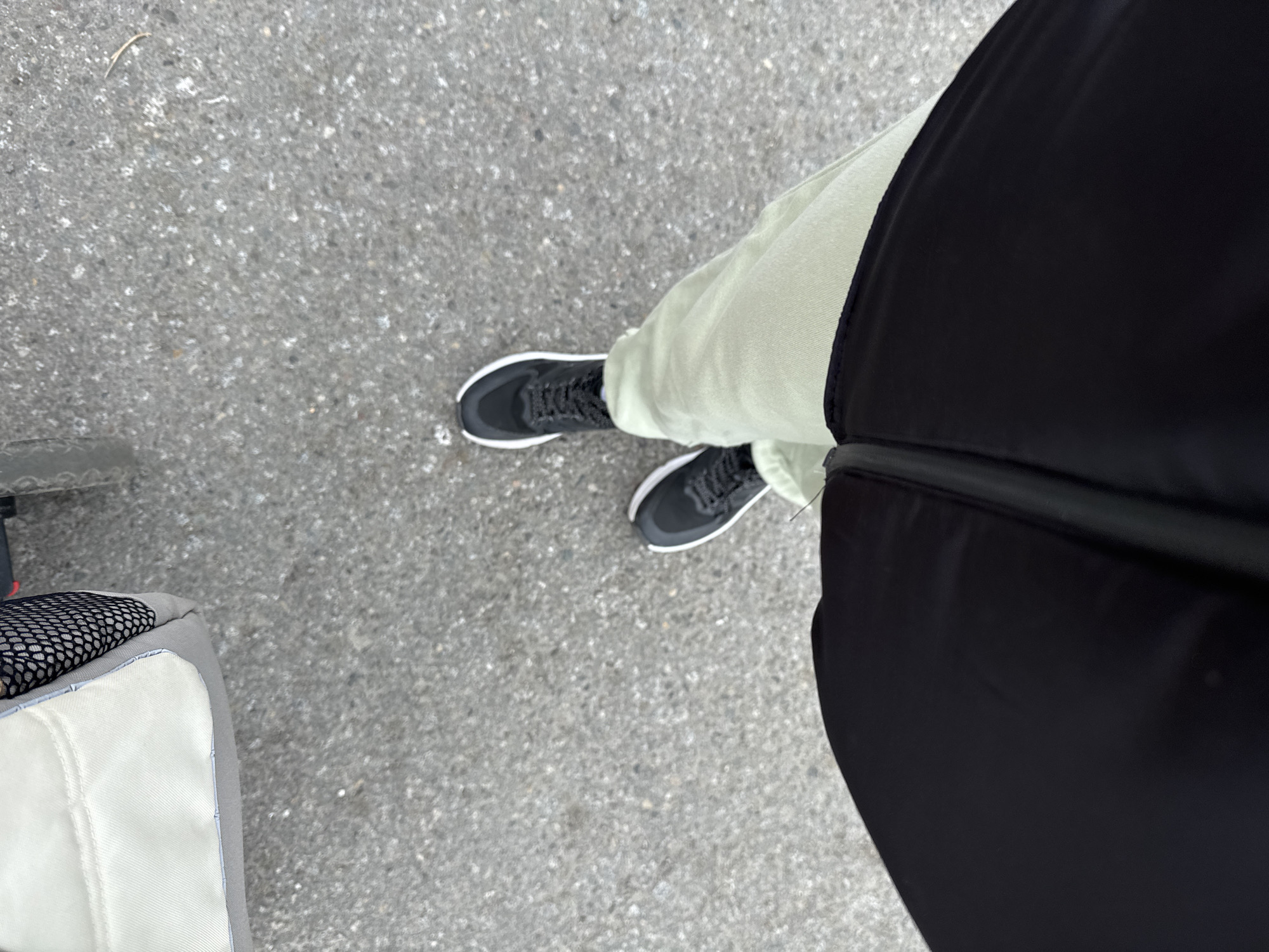 An overhead shot shows a person's legs and feet wearing black athletic shoes and pale green sweatpants. The person is standing on a gray asphalt surface. In the upper left corner of the frame, a portion of a light-colored stroller with a black mesh detail is visible. The scene appears to be outdoors in an urban or suburban setting.