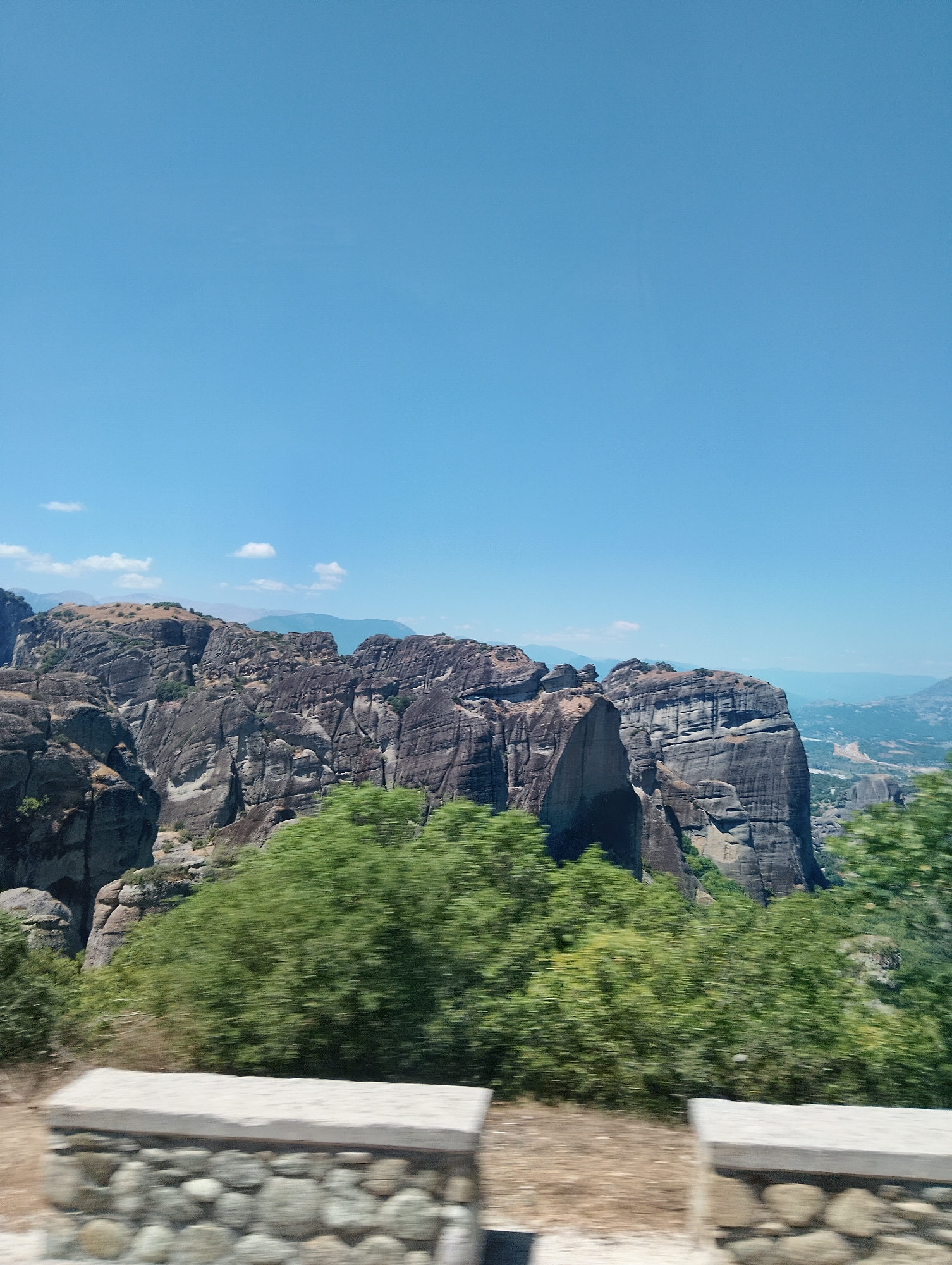 The image shows a panoramic view of the Meteora rock formations in Greece. The foreground features a low stone wall with a gravelly dirt path behind it, partially obscured by blurred green foliage. The midground is dominated by massive, sheer rock cliffs, characteristic of Meteora, with sparse vegetation growing on their ledges and slopes. The background reveals a hazy landscape with rolling hills and a distant valley, all under a clear, bright blue sky with a few wispy white clouds. The overall impression is one of rugged natural beauty and a sense of scale.