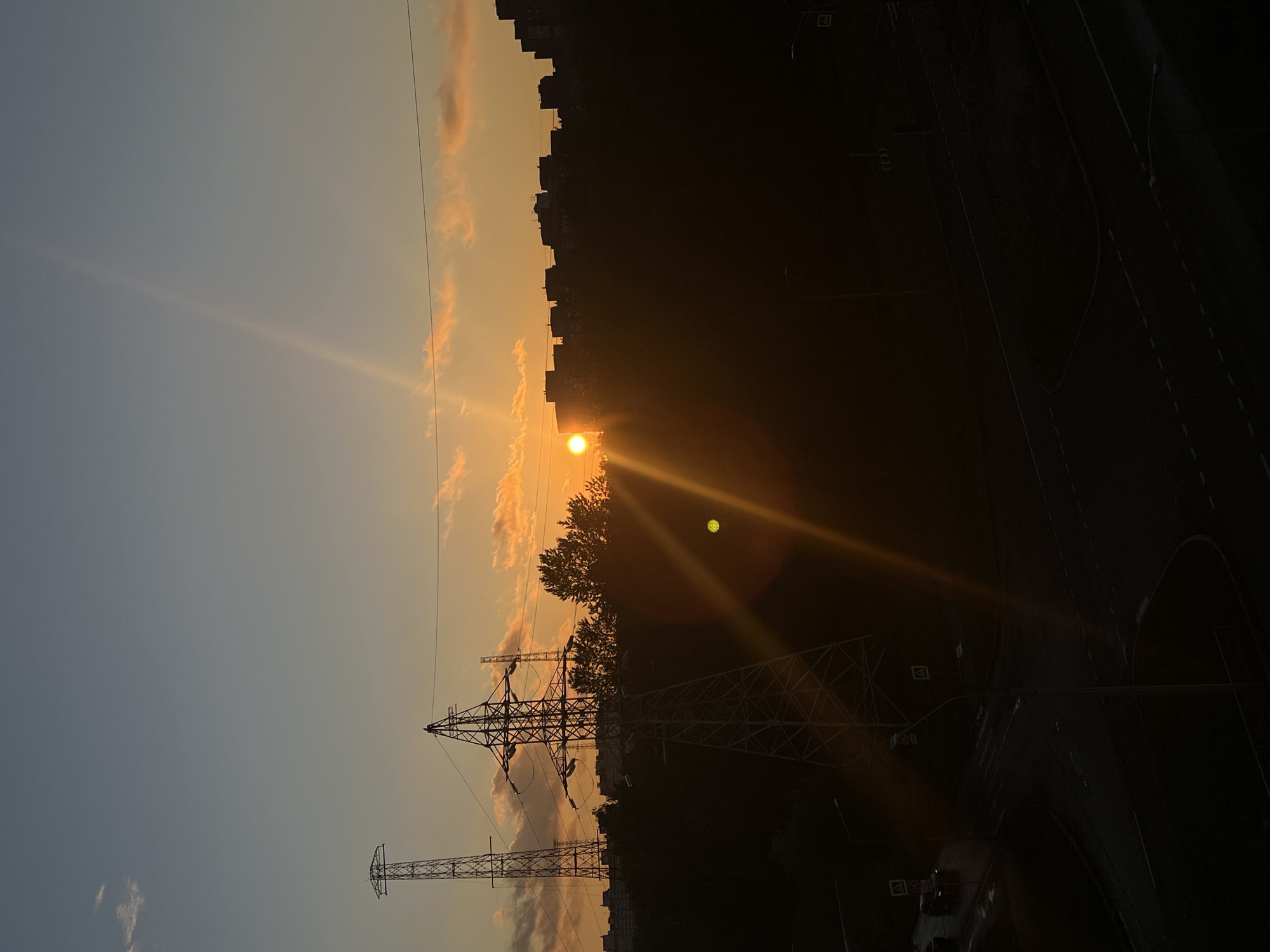 This image depicts a sunset scene in Saint Petersburg, Russia, with an urban landscape. The sun, a bright orange orb, is low on the horizon, casting long rays across the sky and creating a lens flare effect. The sky transitions from a soft blue on the left to a warm orange and pink hue on the right, with a few scattered clouds catching the sunlight.
Prominent in the foreground are two tall electricity pylons, silhouetted against the colorful sky. Their intricate metal structures are clearly visible. Behind the pylons and to the right, the dark silhouettes of urban buildings and trees form a dense backdrop. The building line suggests a densely populated area, likely residential.
In the lower right portion of the image, a dark road network is visible, with streetlights and traffic signs faintly discernible. The overall impression is of a city transitioning from day to night, with the dramatic beauty of the sunset illuminating the urban environment. The time of day is clearly evening, as indicated by the low sun and the beginning of darkness in the cityscape. The weather appears clear, allowing for a vibrant sunset. There is no visible text in the image.