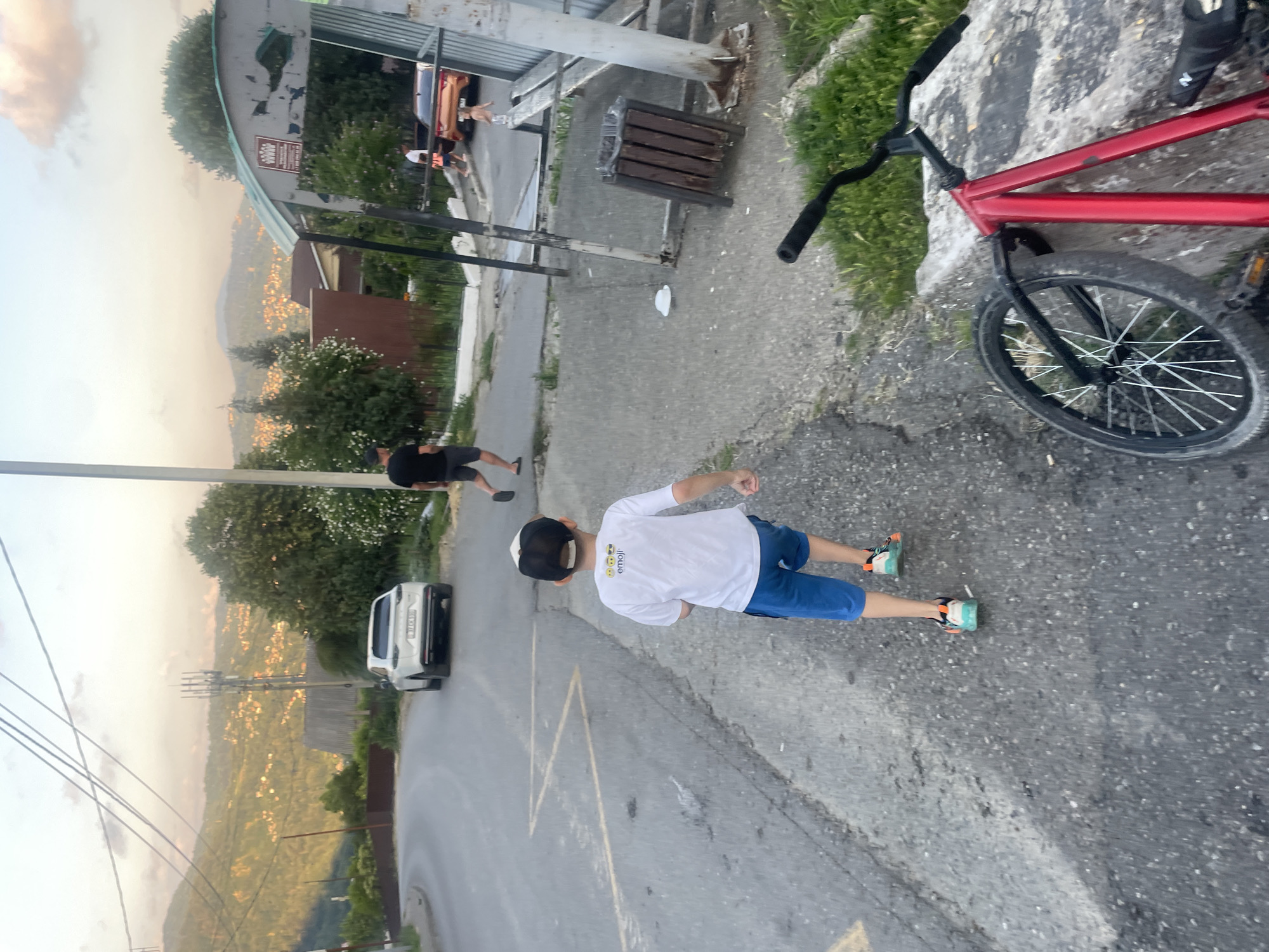 In a scene that appears to be Baranovka, Russia, a young boy in a white t-shirt and blue shorts, wearing a black baseball cap and colorful sneakers, walks away from the camera. He is on a paved road with yellow markings, with a red bicycle to his right. The bicycle has a black handlebar and a black tire with spokes. Behind the boy, a white SUV is parked on the road. Further in the background, a hillside is dotted with houses and trees, and a transmission tower is visible. The sky is bright, suggesting it is daytime, possibly late afternoon or early evening. To the left of the road, there are some structures that look like a bus stop with a bench and a trash can. A man in a black t-shirt and shorts is visible walking away from the bus stop area. The overall mood seems casual and relaxed. The white t-shirt of the boy has some text on the back that appears to be "emoji" followed by smiley faces.