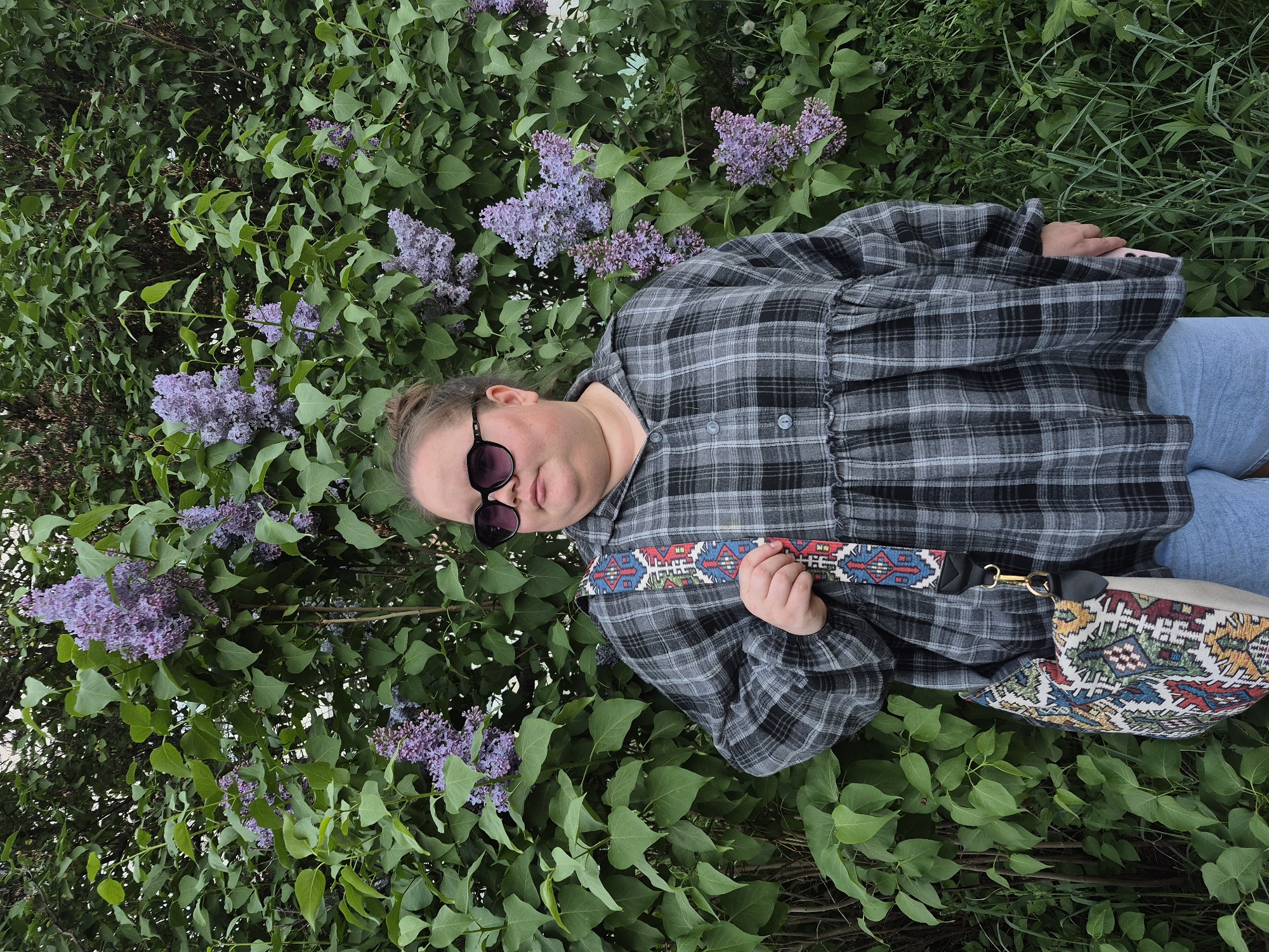 A person is lying amidst a lilac bush. The individual is wearing sunglasses and a gray and black plaid shirt. A patterned bag with a matching strap rests near their hip. The surrounding foliage includes green leaves and clusters of purple lilac blossoms. The location is Chaikovsky, Russia.