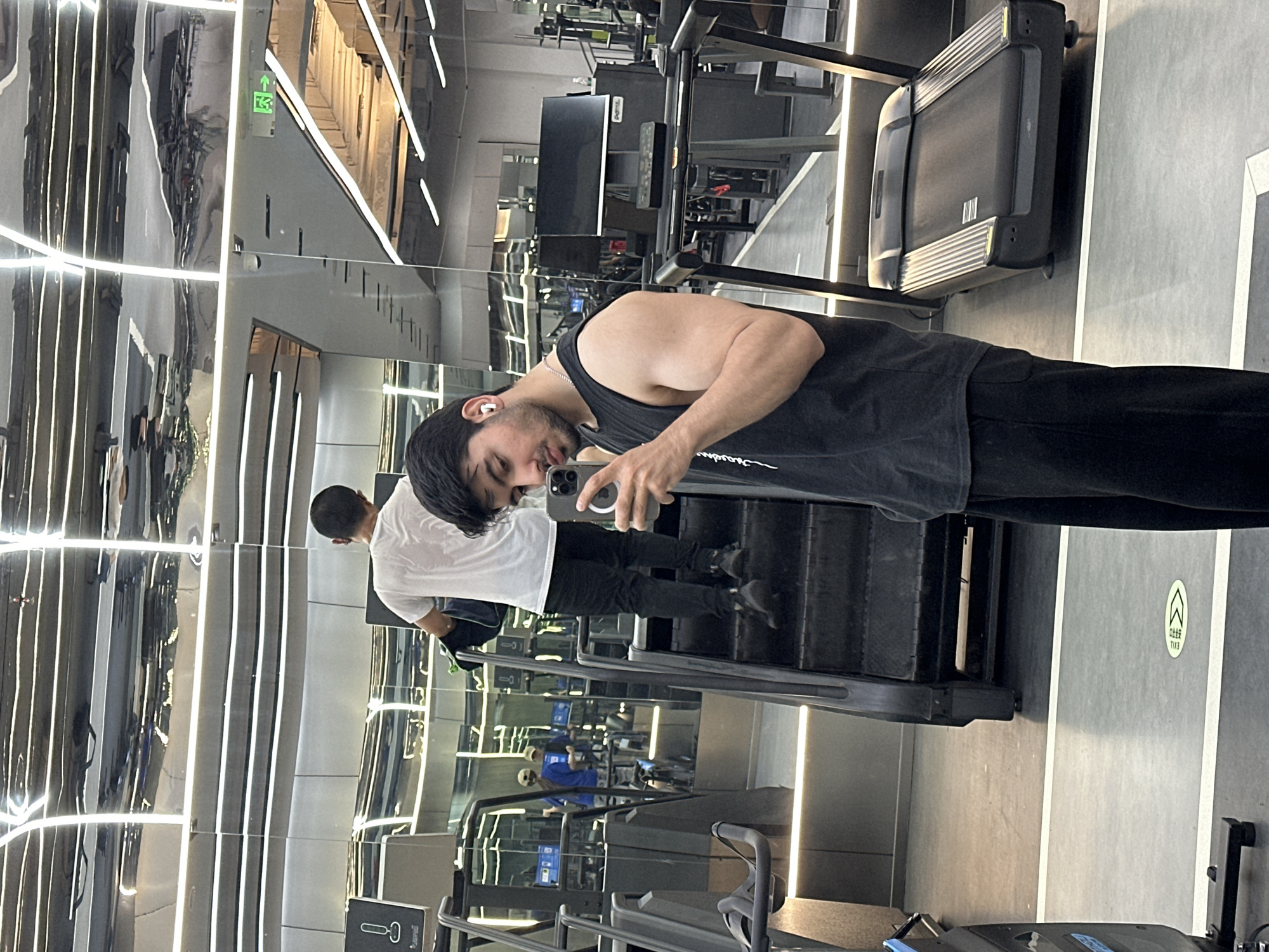The image captures a man taking a selfie in a modern gym with mirrored walls and bright strip lighting, located in Chongqing High-tech Zone, China. The man in the foreground is wearing a black tank top and dark pants, with white earbuds in his ears, and is holding a smartphone to take a picture of his reflection. He has short dark hair and a slight smile.
In the background, multiple reflections of the gym space and another individual are visible due to the extensive mirrors. A second man, reflected behind the selfie-taker, is seen from the back wearing a white t-shirt and dark pants, appearing to be using a stair-stepper exercise machine. Other gym equipment, including treadmills and various exercise machines, are reflected throughout the space.
Notable details include the sleek, metallic gray interior design of the gym. The lighting consists of prominent, long fluorescent strips on the ceiling and walls, creating a bright and clean ambiance.
Visible text includes a green "EXIT" sign with an upward arrow and the Chinese characters "↑ K3 紧急出口" (jǐnjí chūkǒu), meaning "Emergency Exit". Another white oval sign on the wall also shows an upward arrow and the same Chinese characters "紧急出口" (jǐnjí chūkǒu) and "EXIT". The Apple logo is visible on the smartphone held by the foreground man.
