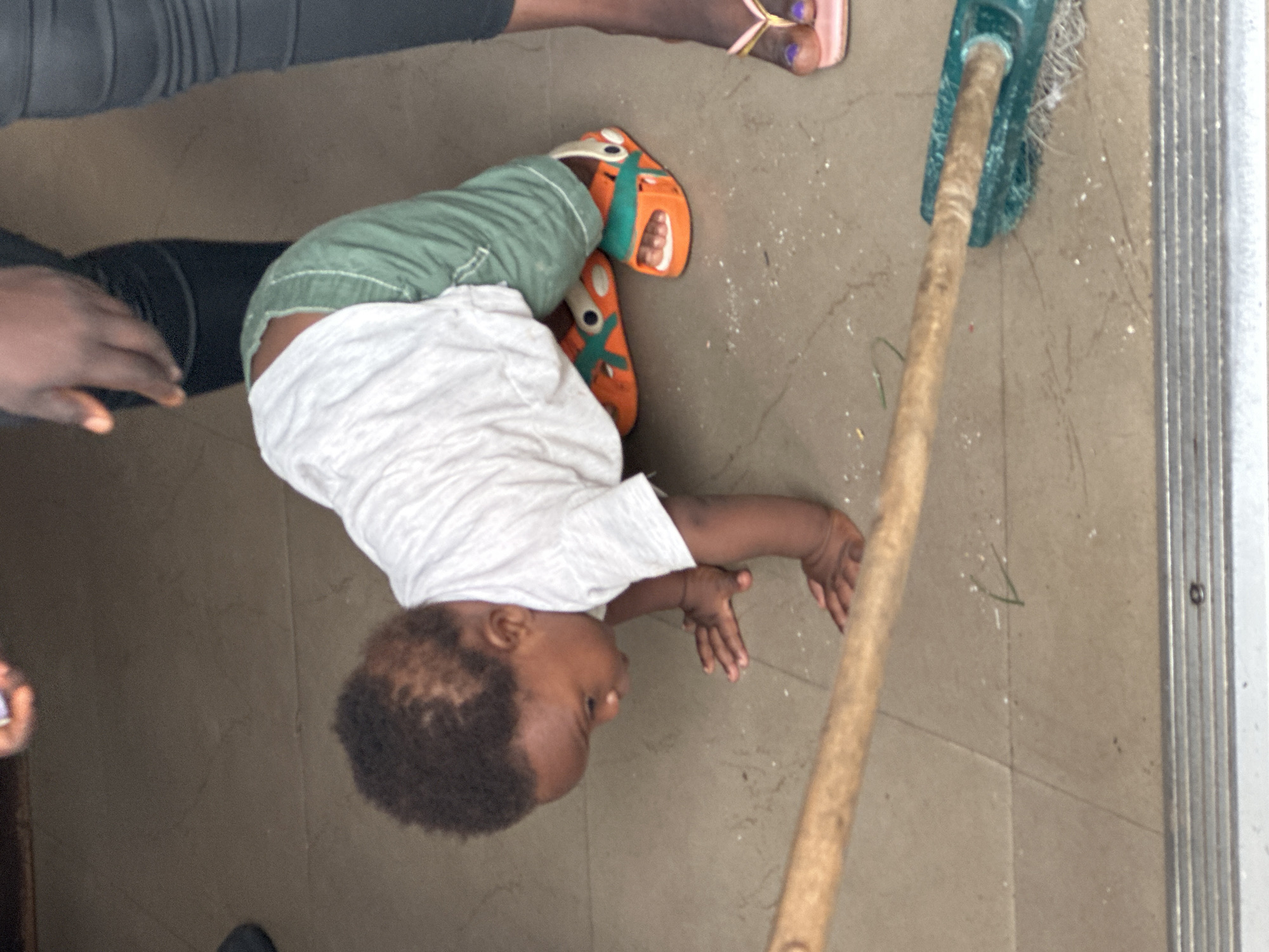 The image shows a young child lying on their back on a tiled floor, with their knees bent and their feet in the air. The child is wearing a white t-shirt and green shorts, and bright orange and green sandals. Their hands are outstretched. To the right of the child, a broom with a blue head and a wooden handle is leaning against the wall. The floor is a neutral-colored tile, with some white specks scattered across it. In the background, the edge of a metal door or frame is visible. Another person's legs are visible in the upper left corner of the image, wearing dark leggings and a dark shoe. A foot wearing a pink flip-flop is visible in the upper right corner. The lighting suggests it is daytime. The scene appears to be indoors, possibly in a home or communal area in Gura Top, Nigeria. The child's posture and outstretched hands might suggest playfulness or an attempt to reach something.