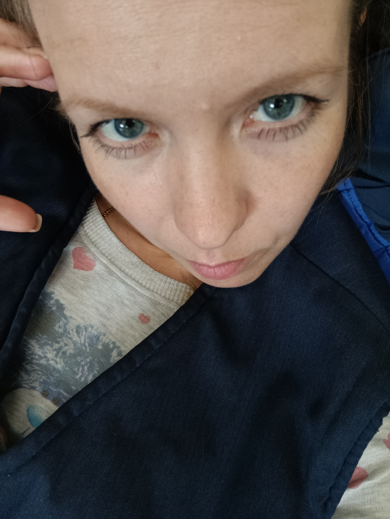 A close-up, high-angle shot of a woman's face. She has fair skin, blue eyes, and dark eyeliner. Her expression appears serious. She is wearing a gray patterned shirt with small pink hearts, covered partially by a dark blue jacket. A fingertip is visible near her forehead. The background shows additional clothing or fabric. There is no information in the image to suggest a location in the city of Chaikovsky, Russia.