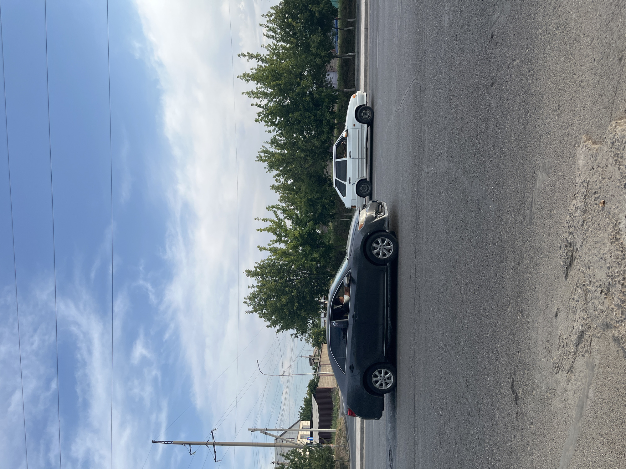In Shymkent, Kazakhstan, on a clear day with scattered clouds, a dark gray sedan is driving on a paved road. The driver, a person wearing a light-colored shirt, is visible inside the car. In the background, a white hatchback is also traveling in the same direction. A line of green trees borders the road, and beyond them, buildings and a power pole with overhead wires are visible. The road surface shows some wear and tear, with cracks and uneven patches.