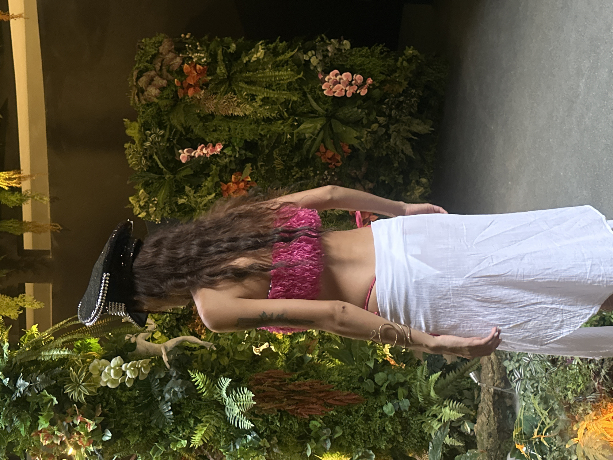 A person is standing in front of a large vertical garden wall composed of various lush green plants and some pink and orange flowers. The person is wearing a black hat with metallic embellishments, a bright pink textured bandeau top, and a long, flowing white skirt. The person's back is to the camera, and they are looking towards the left side of the frame. The lighting suggests an indoor environment, possibly a commercial space or an art installation. The background is dark, highlighting the vibrant green of the garden and the person's attire. The presence of a detailed vertical garden suggests it could be a decorative element in a hotel, restaurant, or event venue in a location like Ko Samui, Thailand, known for its tropical aesthetics.