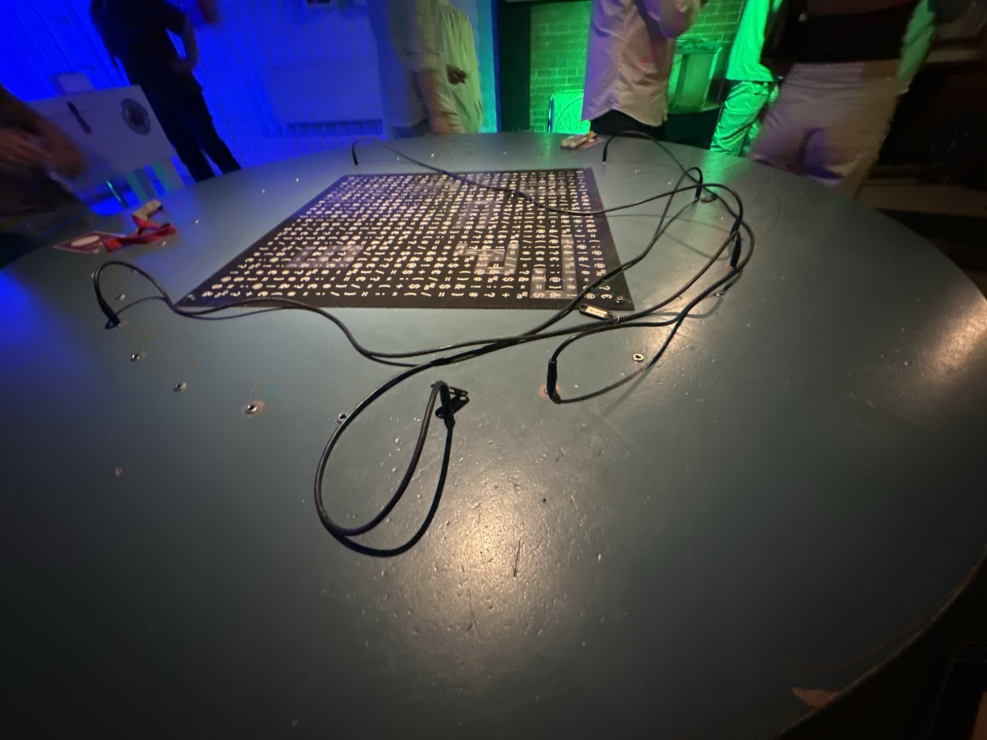 The image depicts a close-up view of a large, round, teal-colored table. A black mat with a dense pattern of white characters, resembling a keyboard layout or code, is centered on the table. Black wires with connectors are spread across the table, originating from the mat and extending outwards. A red lanyard with a white tag is visible on the left side of the table, near the edge. The background is dimly lit, with a bright blue light illuminating the left side of the scene and a green light on the right. Several people are partially visible in the background, their figures blurred and indistinct due to motion or low light. One individual on the left appears to be wearing dark clothing, while others in the background are in lighter colored garments. The overall impression is of an indoor, possibly exhibition or event space, with an interactive or technological display setup. The location is not identifiable from the image alone.