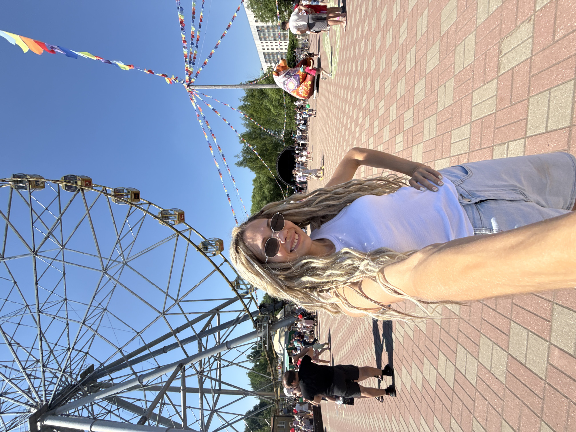 In Заволжье, Russia, a woman with long blonde dreadlocks and sunglasses takes a selfie in an outdoor amusement park. She is wearing a white tank top and light-colored denim shorts. Behind her, a large Ferris wheel dominates the background, with its metal structure against a clear blue sky. Several gondolas are visible on the wheel. Colorful triangular flags are strung across the park, adding a festive atmosphere. In the middle ground, a crowd of people are milling about, and a large, decorated bear-shaped inflatable or art installation is visible to the right, adorned with intricate patterns. Trees and buildings form the backdrop. The ground is paved with red and beige interlocking bricks. The scene suggests a sunny day at a public event or fair.