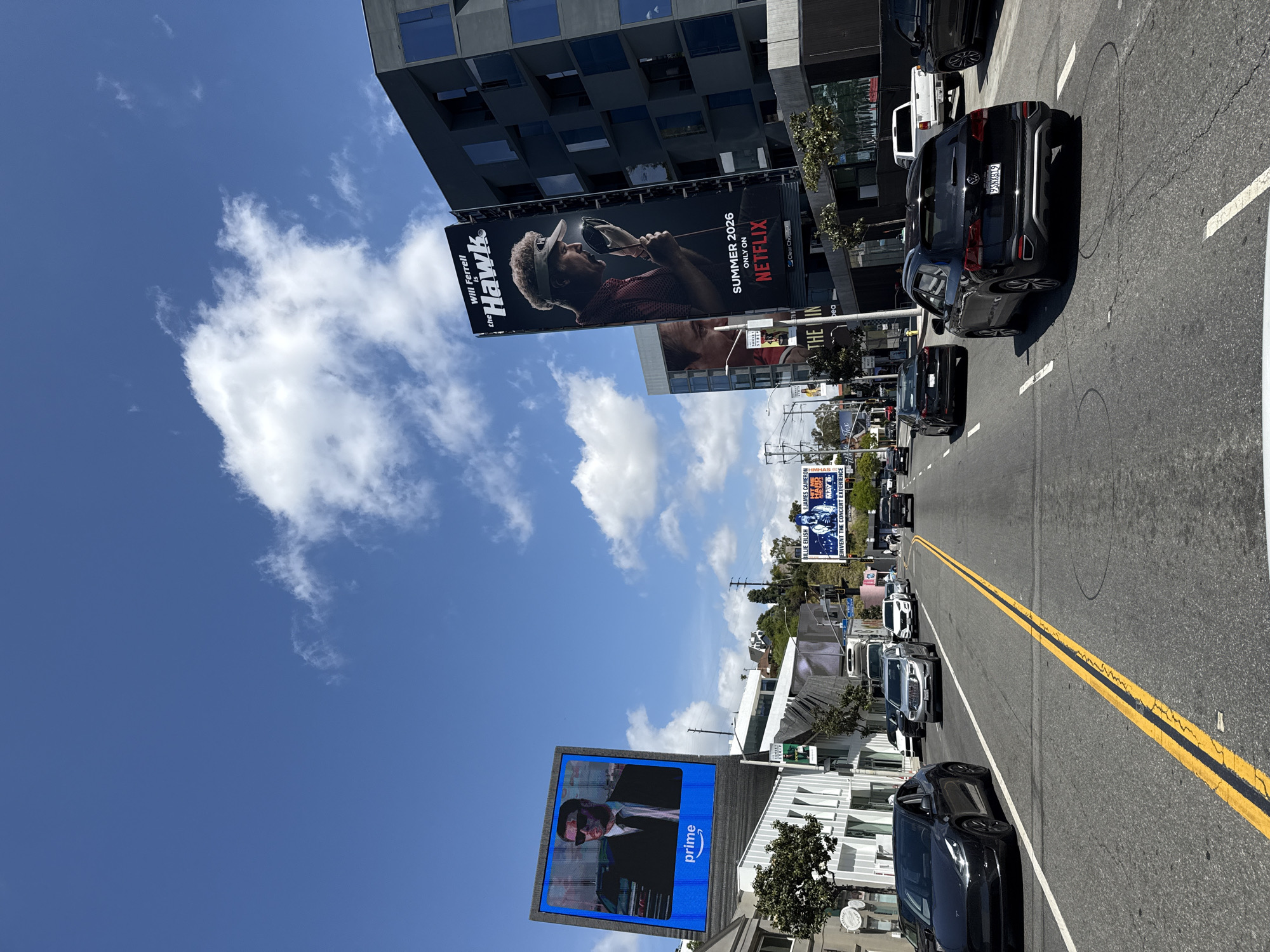 The image captures a street in West Hollywood, United States of America, on a bright day with scattered clouds in a blue sky. Along the roadside, billboards advertise various entities. On one billboard is a poster for the show "Hawk," starring Will Ferrell, which is set to air in the summer of 2026, exclusively on Netflix. Another billboard advertises Prime, with a man in a suit. Furthermore, there is a billboard with Billie Eilish and James Cameron with the text "Reinvent The Concert Experience." The street is lined with cars, some in motion, and the road is divided by double yellow lines. Buildings of varying heights are also visible in the background.