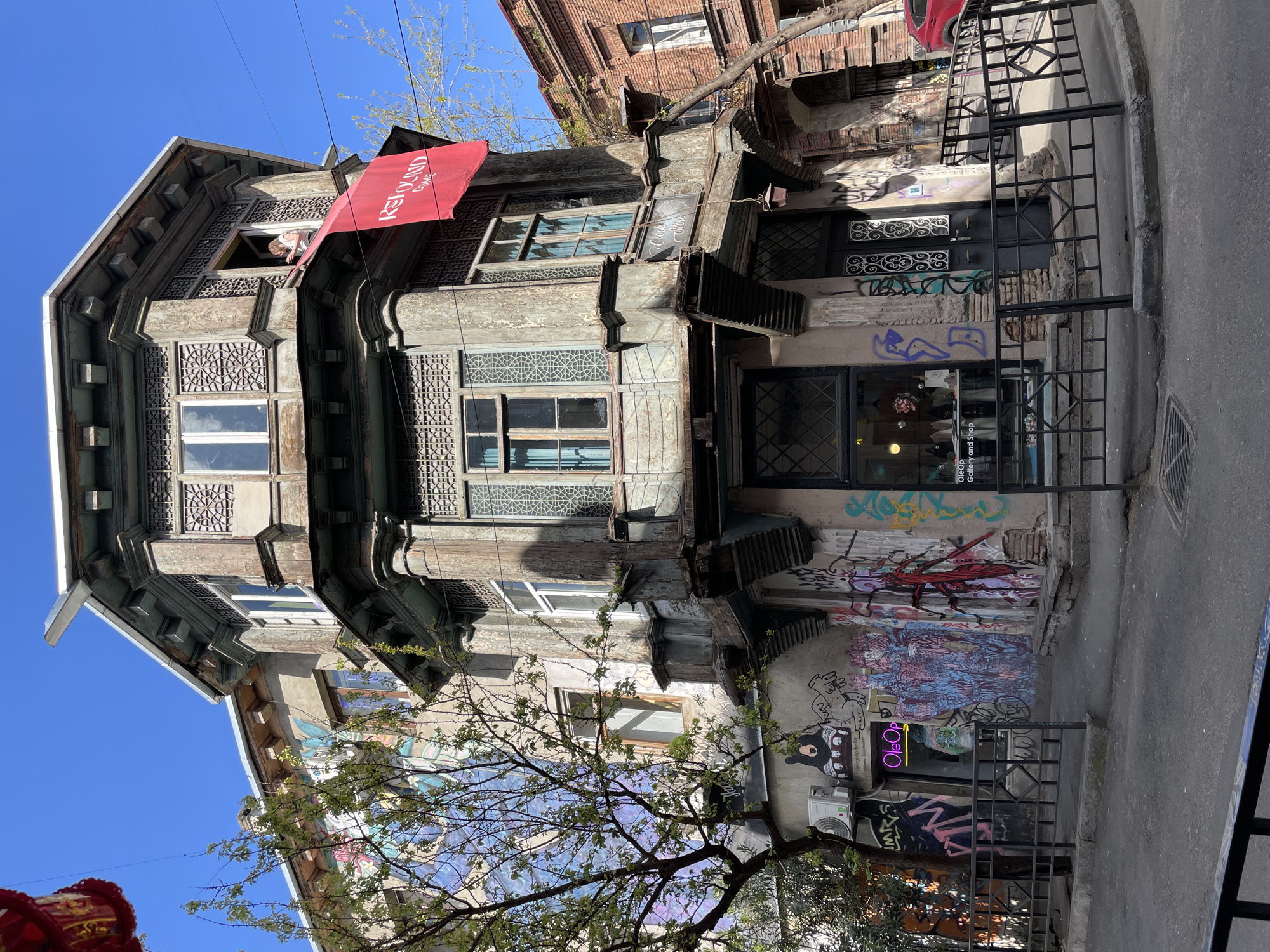 An old, multi-story building with distinctive architecture stands under a clear blue sky in what appears to be an urban setting. The building exhibits signs of age and wear, with peeling paint and weathered wooden elements, yet retains its ornate charm through detailed carvings and patterns around windows and on balconies.
The facade is characterized by intricate geometric patterns on window grilles and decorative wooden panels. On the top floor, a person is seen peering out of an open window. A red banner or flag, bearing the white text "RESOUND CLUB," hangs from an upper balcony, partially obscuring the details of that section.
The lower levels of the building are heavily covered in colorful graffiti, featuring various tags, abstract designs, and a discernible cartoonish bear-like figure on the ground floor. Several businesses operate on the street level; one prominent sign reads "OleOp Gallery and Shop" with "OleOp" illuminated in neon purple. Another sign above a doorway indicates "Cafe & Wine." The building's entrance and ground-floor windows are secured by black metal railings and gates.
In the foreground, a deciduous tree with fresh green leaves partially frames the scene. Overhead wires are visible against the bright sky. The street and sidewalk are paved with gray asphalt and concrete. The bright daylight suggests a sunny day, casting some shadows. The overall scene captures a blend of historic architecture, urban decay, and contemporary street art, characteristic of older city districts. The location context provided is Tbilisi, Georgia.