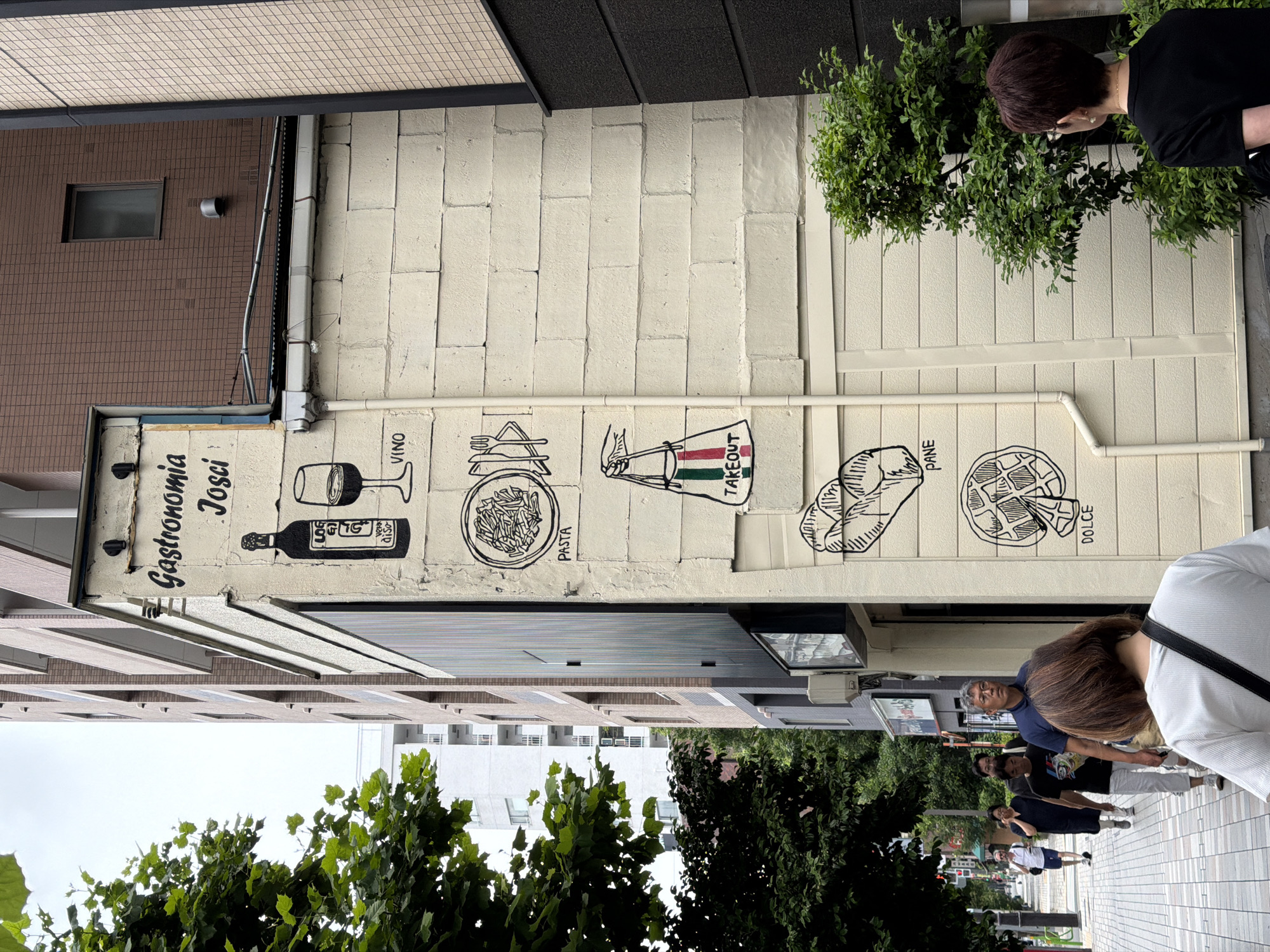 The image shows the exterior of a building with a mural painted on its side. The mural depicts a wine bottle and glass, a plate of pasta with utensils, a shopping bag with an Italian flag design, a loaf of bread, and a waffle, all labeled with words like "Vino," "Pasta," "Takeout," "Pane," and "Dolce." The building appears to be a restaurant or shop located in the city of Chuo, Japan, as indicated by the context of the provided information. The mural is rendered in a simple black and white line-drawing style. In the background, there are other buildings, trees, and a sidewalk where several people are walking. The sky is overcast.
