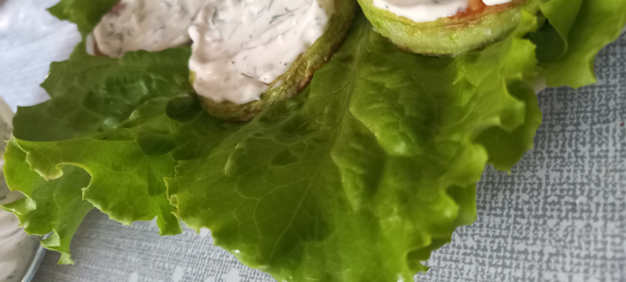 This image captures a close-up view of food presented on a patterned surface, located in Kryvyi Rih, Ukraine. The central subject is a vibrant green lettuce leaf, exhibiting pronounced venation and a crinkled texture, serving as a base. Resting on the lettuce are multiple circular slices, consistent with cooked zucchini or squash, characterized by a light green interior and subtly browned edges. Each slice is topped with a generous portion of a viscous, off-white, creamy sauce. Small, dark green particulates, consistent with chopped dill or similar herbs, are suspended within the sauce. The supporting surface is light gray with a repeating, irregular darker gray pattern. A portion of an out-of-focus white container or dish is visible in the upper left background. No individuals or overt actions are depicted.