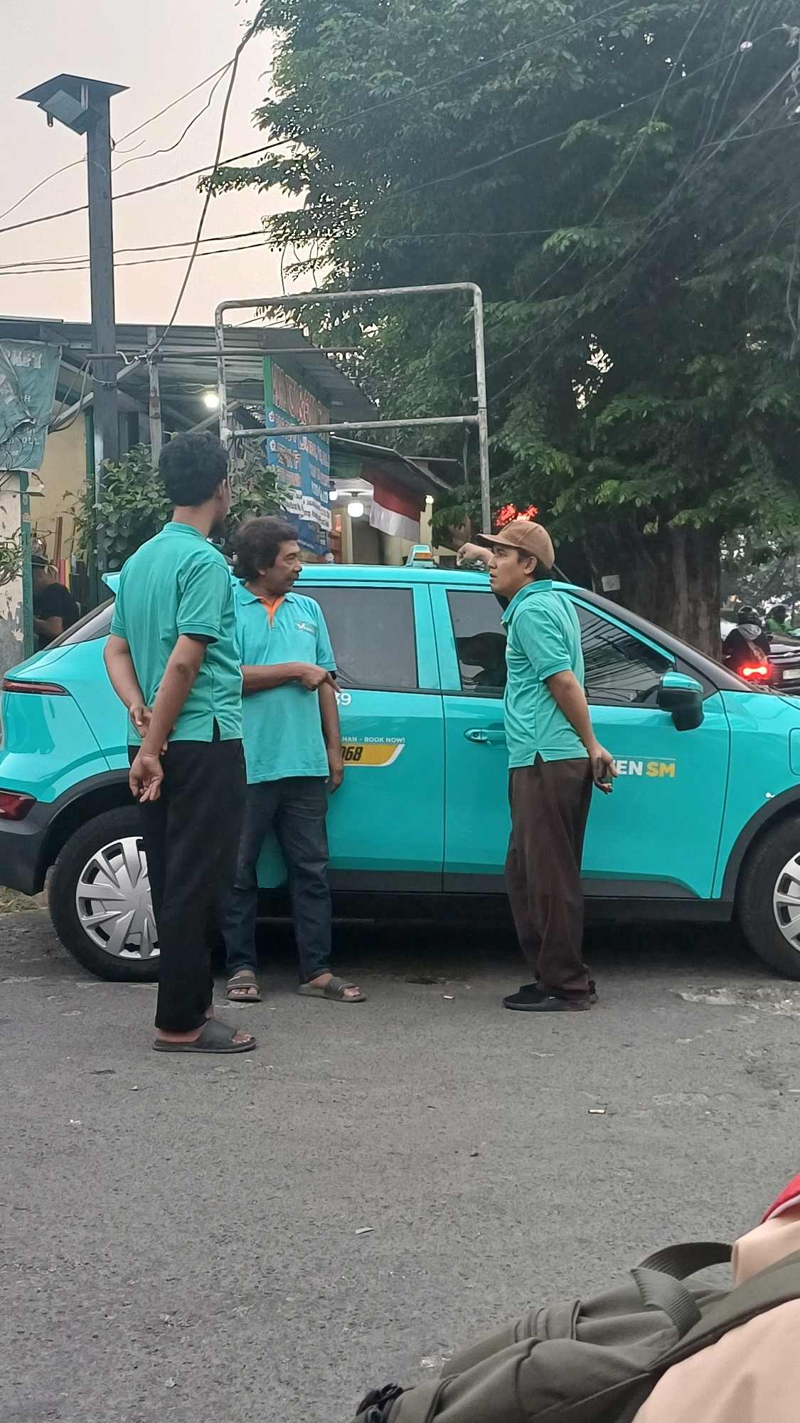 In South Jakarta, Indonesia, three men in teal shirts stand near a teal-colored car. One man, wearing a brown cap, gestures with his right hand while facing the car. Another man in a teal shirt and blue jeans points towards the car with his left hand. The third man, also in a teal shirt and black pants, stands with his back to the camera, facing the other two. The car has markings that say "AKU - BOOK NOW!" and "468" along with "SM". The scene appears to be on a street with trees and a lamppost visible in the background. The sky is overcast.