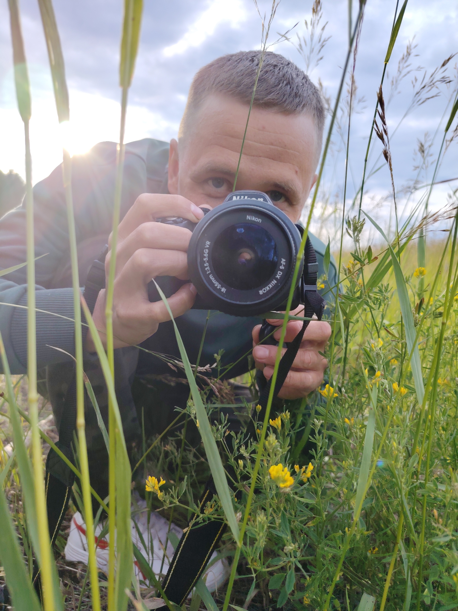 A man is shown from the chest up, holding a black Nikon DSLR camera with a black strap. He is crouching in tall green grass with small yellow flowers, with the sun shining brightly behind him and to his left. The man has short brown hair and is wearing a dark green hooded jacket. His eyes are focused on the camera, suggesting he is taking a photo. The scene appears to be outdoors, possibly in a field or meadow, during daylight. The sky is visible behind him, with some clouds. White sneakers with red accents are visible at the bottom of the image, peeking through the grass. Text on the camera lens reads "NIKON AF-S DX NIKKOR 18-55mm 1:3.5-5.6G VR". The overall mood seems natural and candid, capturing a moment of photography in an outdoor setting.