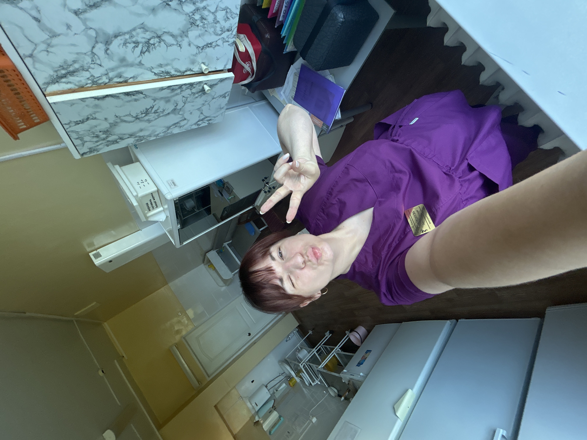 A young woman wearing a purple medical uniform with an identification badge poses for a selfie in a room that appears to be a medical facility or laboratory, possibly located in the city of Фоки, Russia. The room contains several white refrigerators, shelves with medical equipment, and a sink. The woman is making a peace sign with her right hand and puckering her lips. The overall lighting suggests it is daytime, with natural light entering from the left side of the frame.