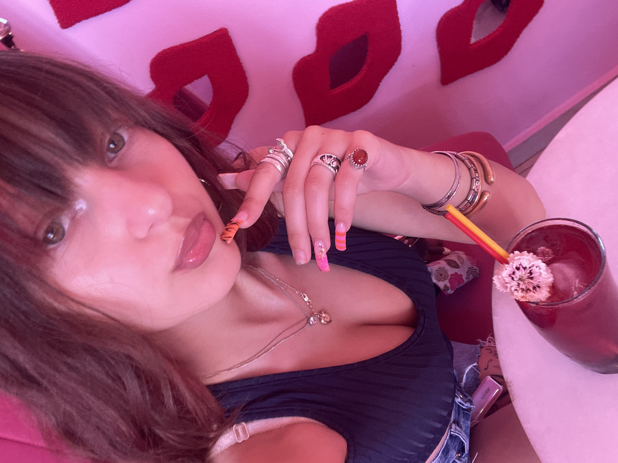 The image shows a close-up, selfie-style shot of a young woman with long, dark brown hair and bangs. She is wearing a dark, ribbed tank top and denim shorts. Her left hand, adorned with colorful, tiger-striped and pink-tipped nails and several rings, is raised near her face. She is holding a red drink with a yellow and red straw, topped with a small white flower. The background is pink and features large, red, stylized lips. The overall aesthetic suggests a vibrant and playful atmosphere, possibly a bar or cafe in Paphos Municipality, Cyprus, known for its stylish and themed establishments.