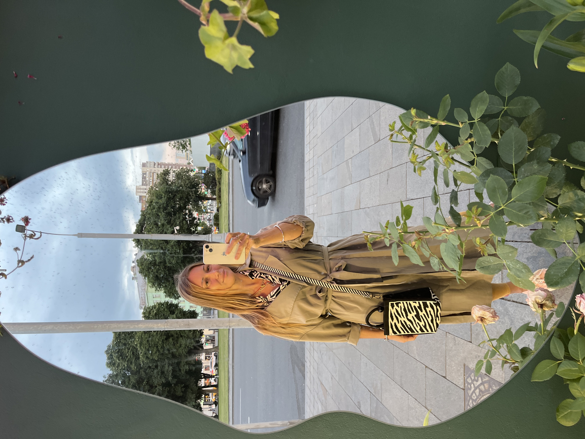 In this mirror reflection, a woman stands outside, holding a smartphone to take a selfie. She has blonde hair and is wearing a tan trench coat over a black and white zebra-print top. A small black purse with a zebra-print design is slung across her body. The setting appears to be an urban environment, possibly a park or a sidewalk, with a street and buildings visible in the background. Trees line the street, and the sky is overcast, suggesting it might be a cloudy day. A dark-colored car is parked on the side of the road. The reflection also captures some of the surrounding greenery, including a branch with leaves and pink flowers, and another plant with lush green leaves. The overall impression is that of a casual outdoor snapshot. The image is oriented vertically, with the reflection appearing upside down relative to the framing of the mirror.