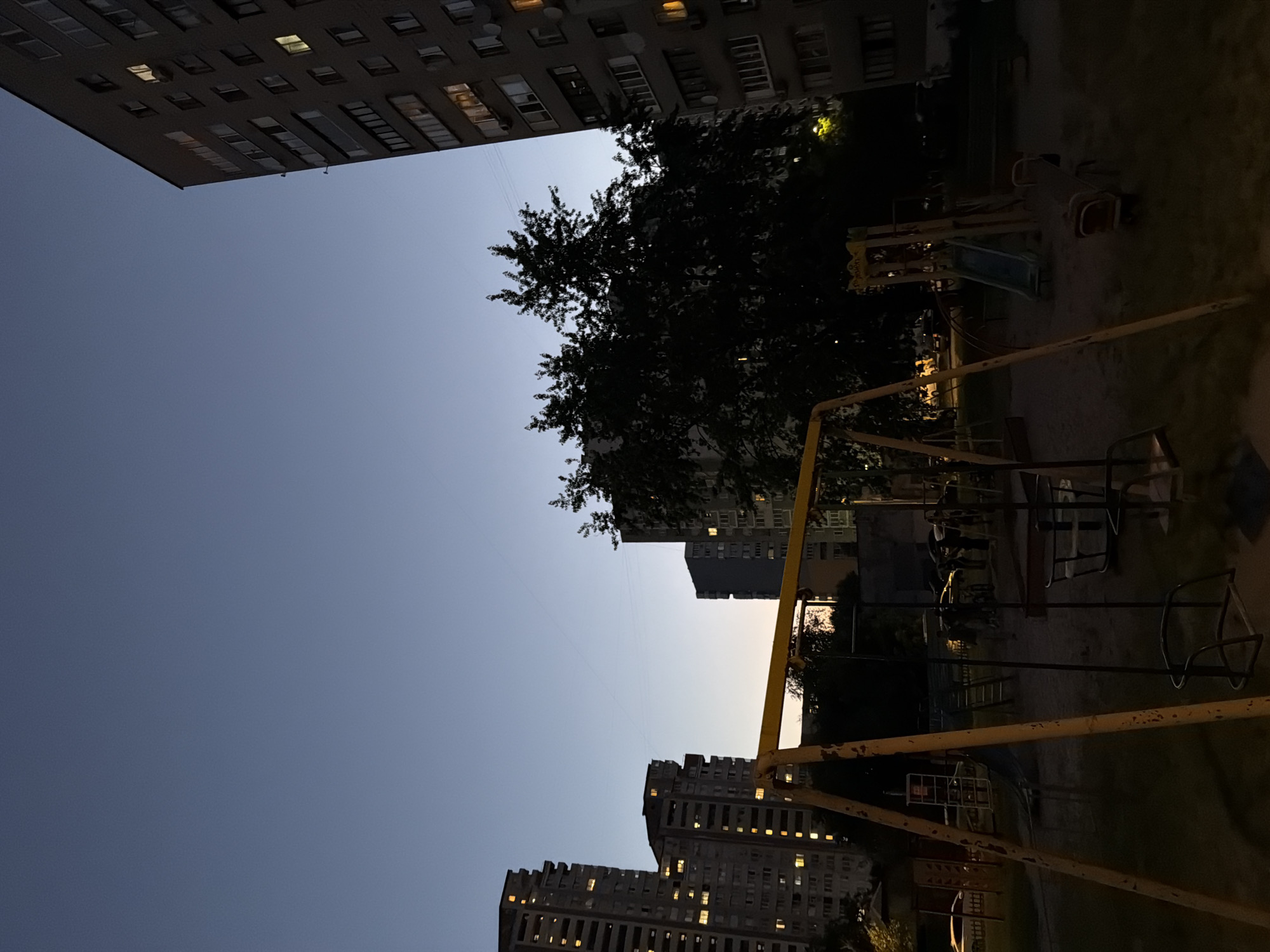 The image depicts a dusk scene in a residential courtyard in Kyiv, Ukraine. The sky is a deep twilight blue, transitioning to a lighter hue near the horizon. Several multi-story apartment buildings, characteristic of Soviet-era architecture, are visible, their windows illuminated with warm yellow light. These buildings are a prominent feature of the urban landscape.
In the foreground, a playground is set up. A yellow metal swing set with an empty swing is positioned prominently. Other playground equipment, including a slide and a seesaw, are also present, although partially obscured by shadows and the surrounding darkness. The ground of the playground appears to be sand or dirt. Trees with dark, silhouetted foliage rise behind the playground, adding to the evening atmosphere. In the background, the faint outlines of more apartment buildings and parked cars can be discerned. The overall impression is one of a typical, everyday urban environment at the end of the day.