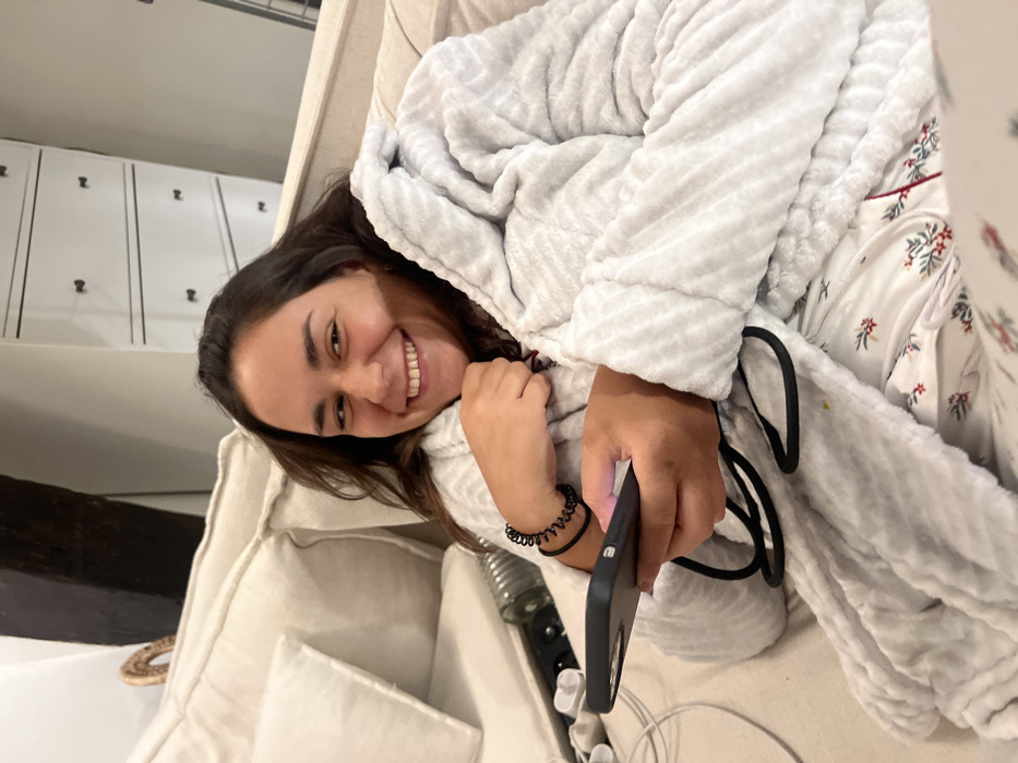 A young woman in a hooded gray robe and printed pajamas smiles at the camera while reclining on a white sofa. She holds a black smartphone in her hand, a black coiled hair tie on her wrist. A water bottle and tangled white charging cables sit nearby. A dark beam and white cabinets are visible in the background.