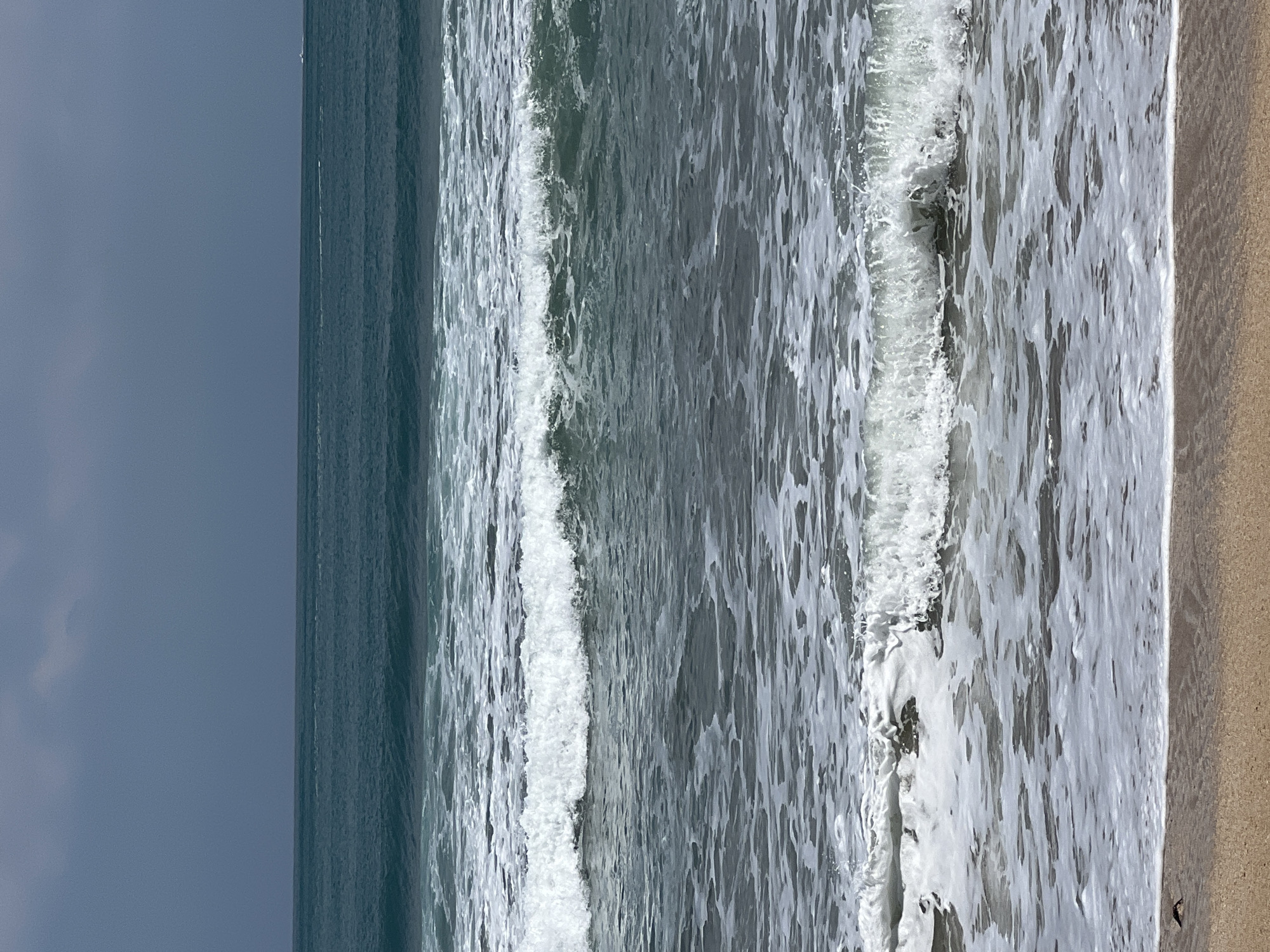 The image depicts a serene seascape under an overcast sky. The ocean is a deep teal, with visible waves rolling towards the shore. Several waves are captured mid-crest, showcasing frothy white caps and the translucent green of the water. The foreground is a sandy beach, appearing wet and smooth near the waterline. A single small dark object, possibly a bird or a piece of debris, is visible on the sand near the left edge of the frame. The horizon line is sharp, separating the vast ocean from the cloudy sky. There are no people or explicit man-made structures visible in the image. The scene is devoid of any specific indicators that would place it in Kuta, Indonesia.