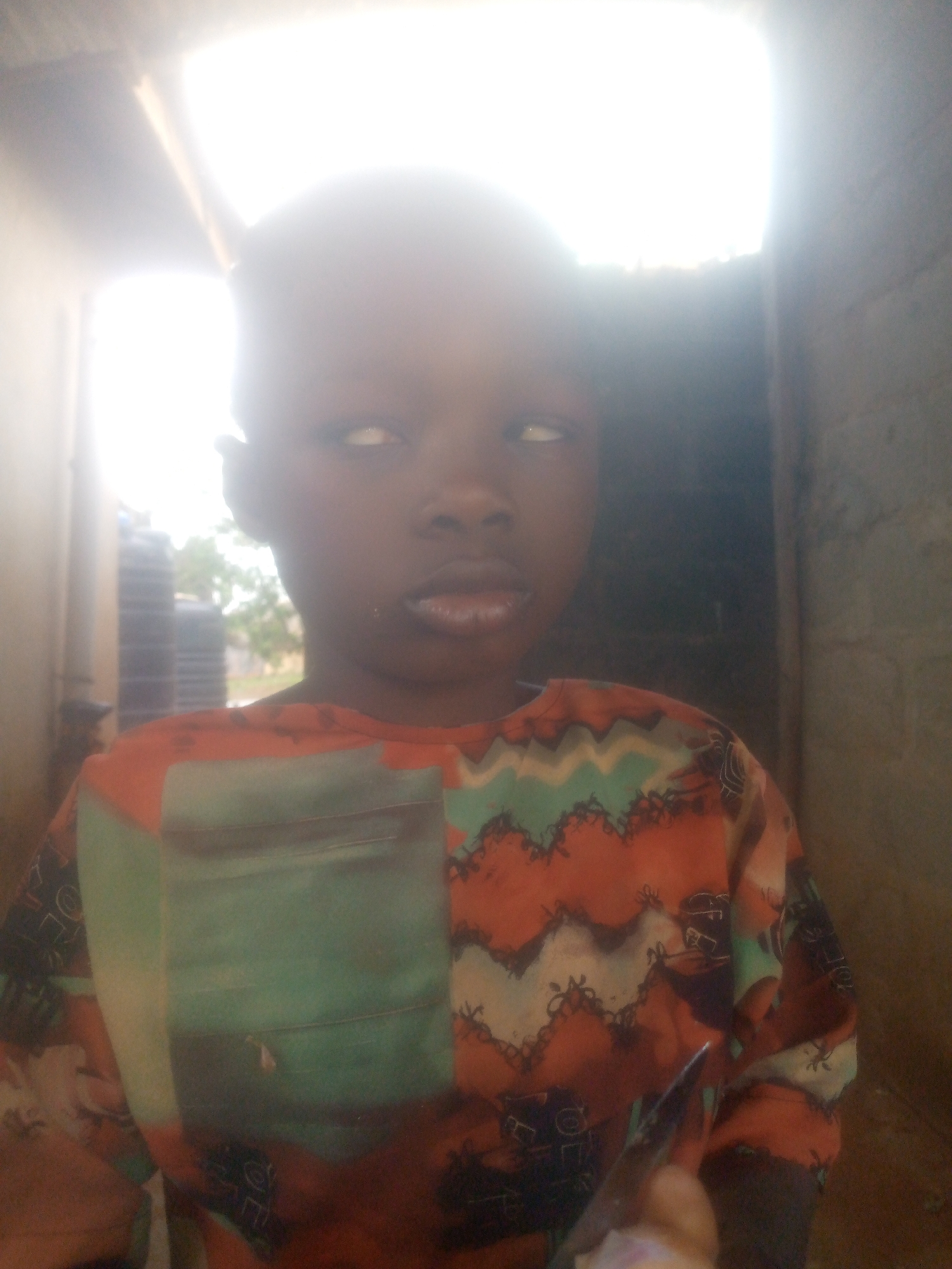 The photograph depicts a young person with dark skin, likely a child, standing indoors, possibly near a doorway or opening that is emitting a strong light. The child is wearing a patterned shirt with shades of orange, green, and black. The eyes appear to be looking upwards and slightly to the left. The surrounding environment is somewhat dark, with details of a wall or structure visible in the background. The location is Unknown, Unknown.