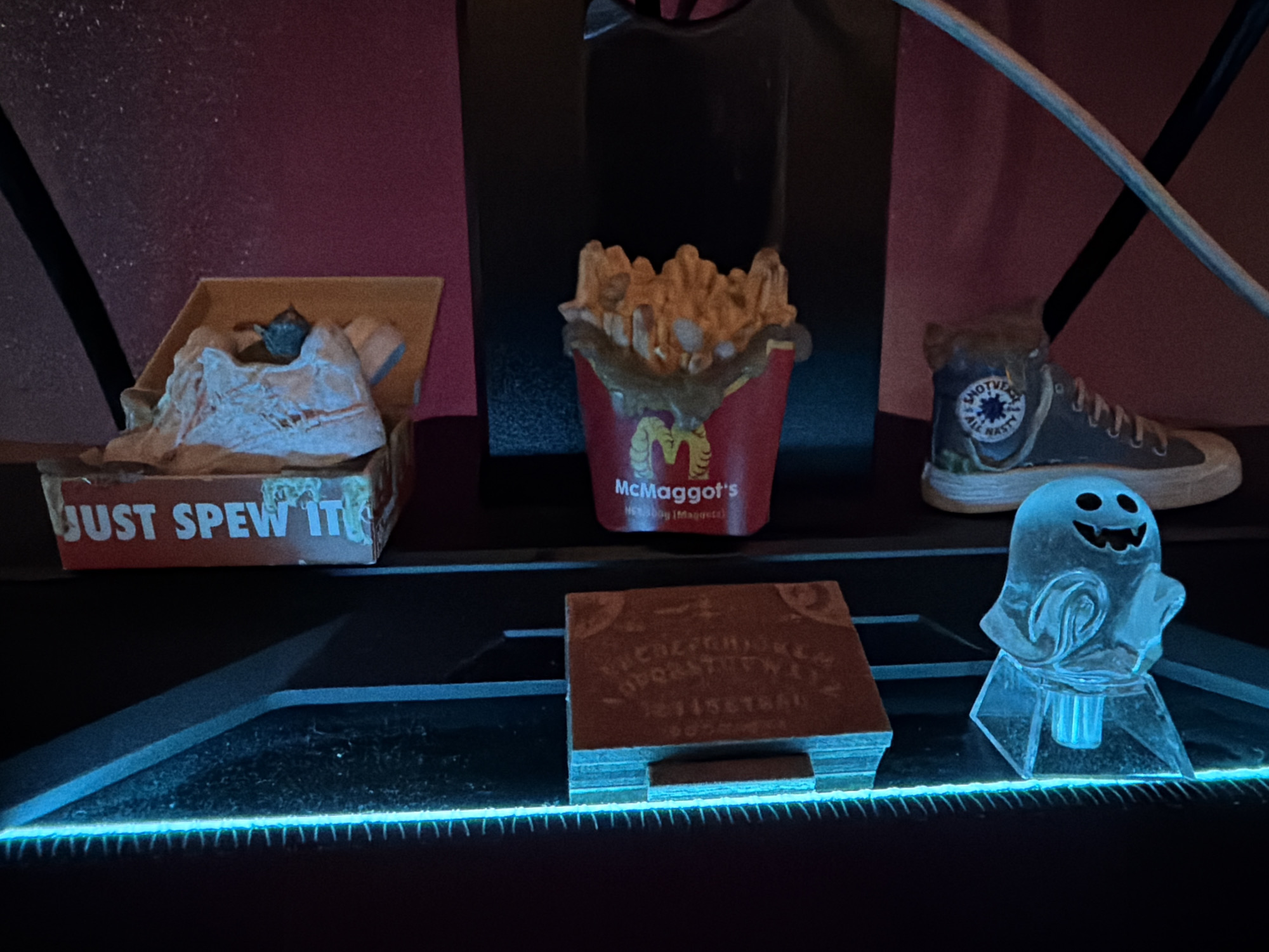 The image shows a shelf with novelty items. From left to right, there is a box labeled "JUST SPEW IT" containing a sneaker, a container of fries labeled "McMaggots," a high-top sneaker with a patch reading "NOT VERY ALL NASTY," a planchette for a Ouija board, and a small, translucent ghost figure. The items are arranged on a dark shelf that is illuminated with a blue light strip along the front edge. The background is dark and partially obscured, with hints of pink and black. Location is unknown.