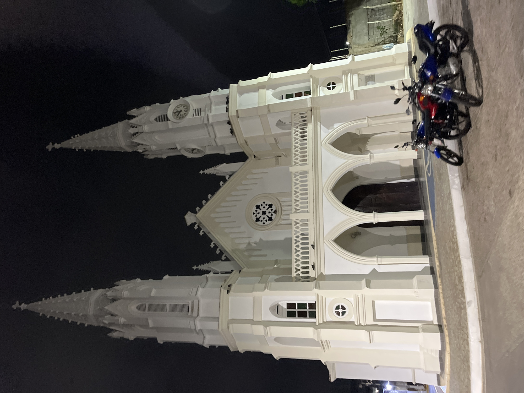 The image captures the Iglesia San Juan Evangelista, a white church in Juan Griego, Venezuela, at night. The church features two prominent spires, a clock tower, and arched doorways. Two motorcycles are parked on the sidewalk in front of the building. The sky is dark.