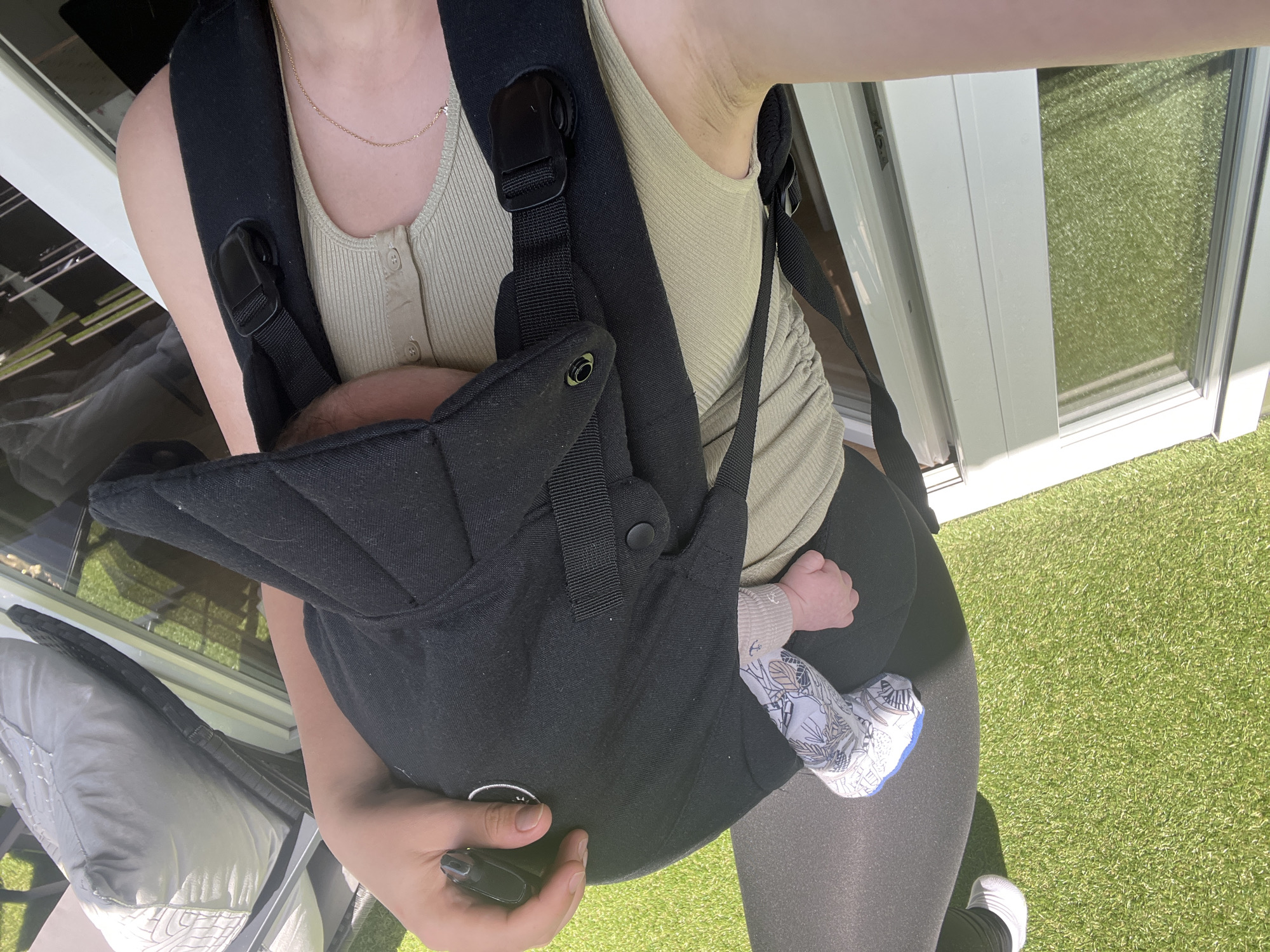 A person is holding a baby in a black carrier, outdoors on a sunny day. The person's right hand is visible, holding a black object, possibly a phone. The baby's left hand and leg are also visible, with the baby wearing a light-colored outfit and patterned leggings. The background shows a grassy area and a white structure, potentially part of a house or building. The image is taken from a high angle, looking down at the person and baby. The location is not identifiable as Garmisch-Partenkirchen, Germany.