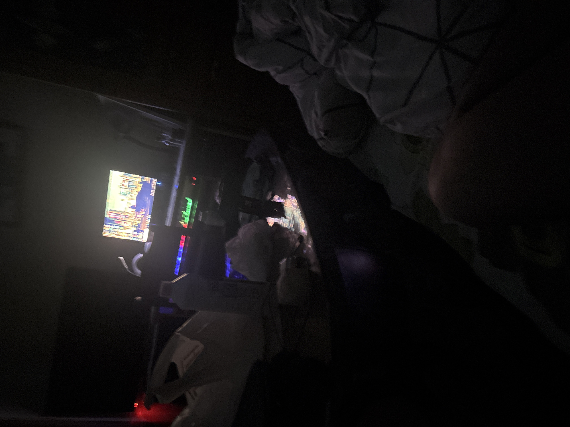 The scene appears to be inside a darkened room. In the left portion of the image, a brightly lit computer monitor displays an image resembling a cityscape, possibly a video game or application interface. To the right of the monitor are components of a computer system or gaming setup, featuring lights and potentially electronic peripherals. On the right side of the image, a dark figure that appears to be a person is partially visible, partially covered by a patterned duvet. The environment is dimly lit, making detailed observation of objects and interactions challenging. The image lacks identifiable landmarks or features that would indicate it is taken in Yerevan, Armenia.