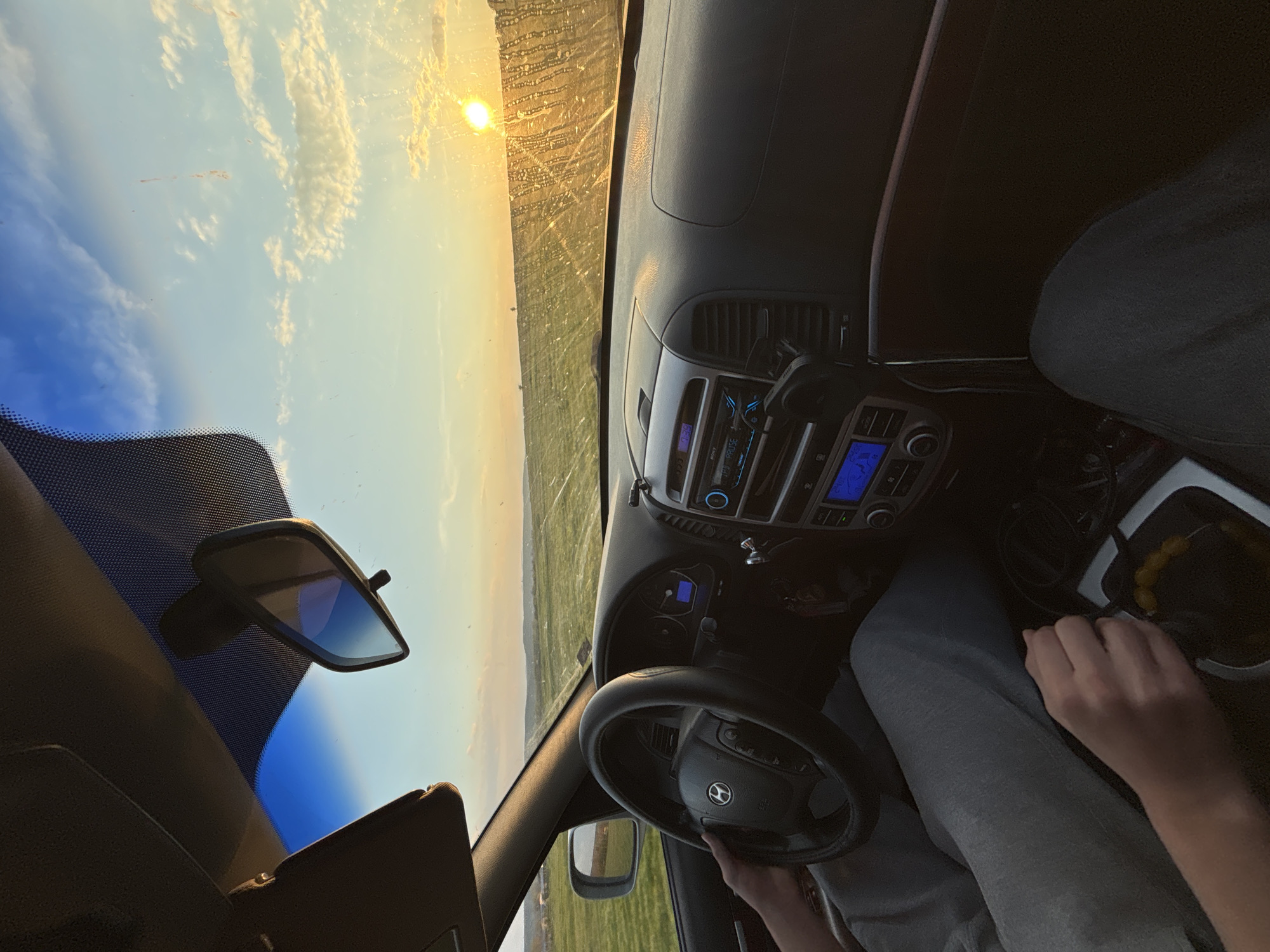 A first-person perspective shot from inside a car. The dashboard, steering wheel, and rearview mirror are visible. A person is driving, hands on the steering wheel. The exterior shows a rural landscape with a field and a partly cloudy sky. The sun is visible in the distance. The location is the city of Lehchevo, Bulgaria.
