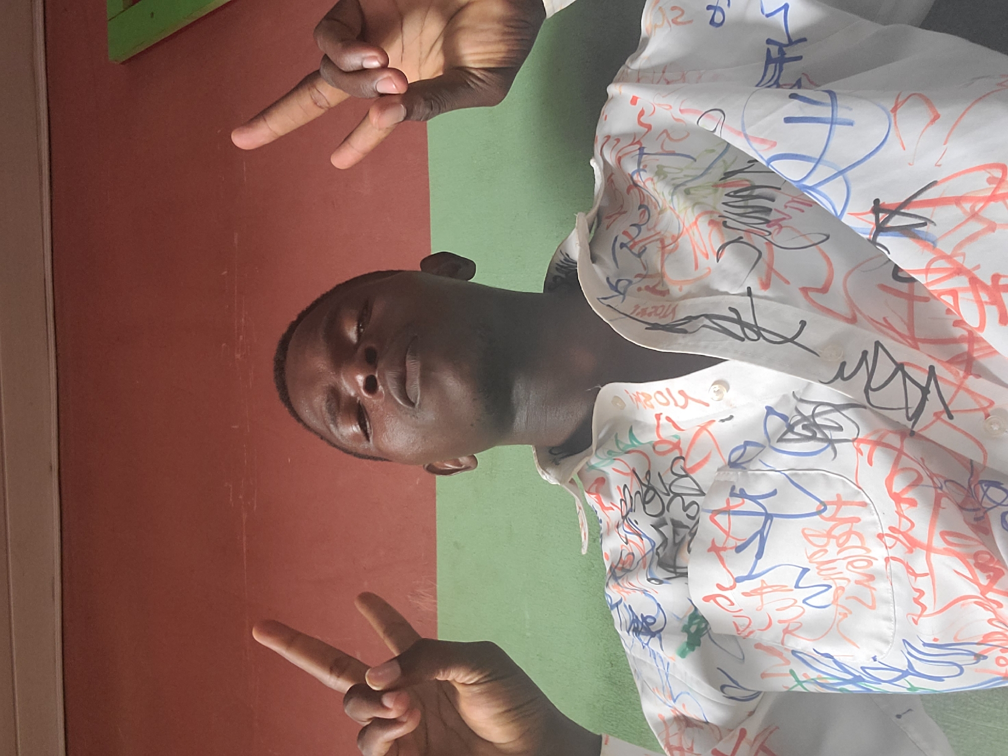 A low-angle, medium shot shows a Black male with short hair and dark skin, looking up and making a peace sign with both hands. He is wearing a white button-up shirt covered in colorful graffiti-style scribbles. The man's face is partially obscured by the angle of the shot. He has his eyes closed and his lips are slightly parted. The background is a two-toned wall, with a reddish-brown color on the top half and a light green color on the bottom half. There are no identifiable landmarks indicating the city of Gura Top, Nigeria, or any specific urban setting. The image appears to be taken indoors.