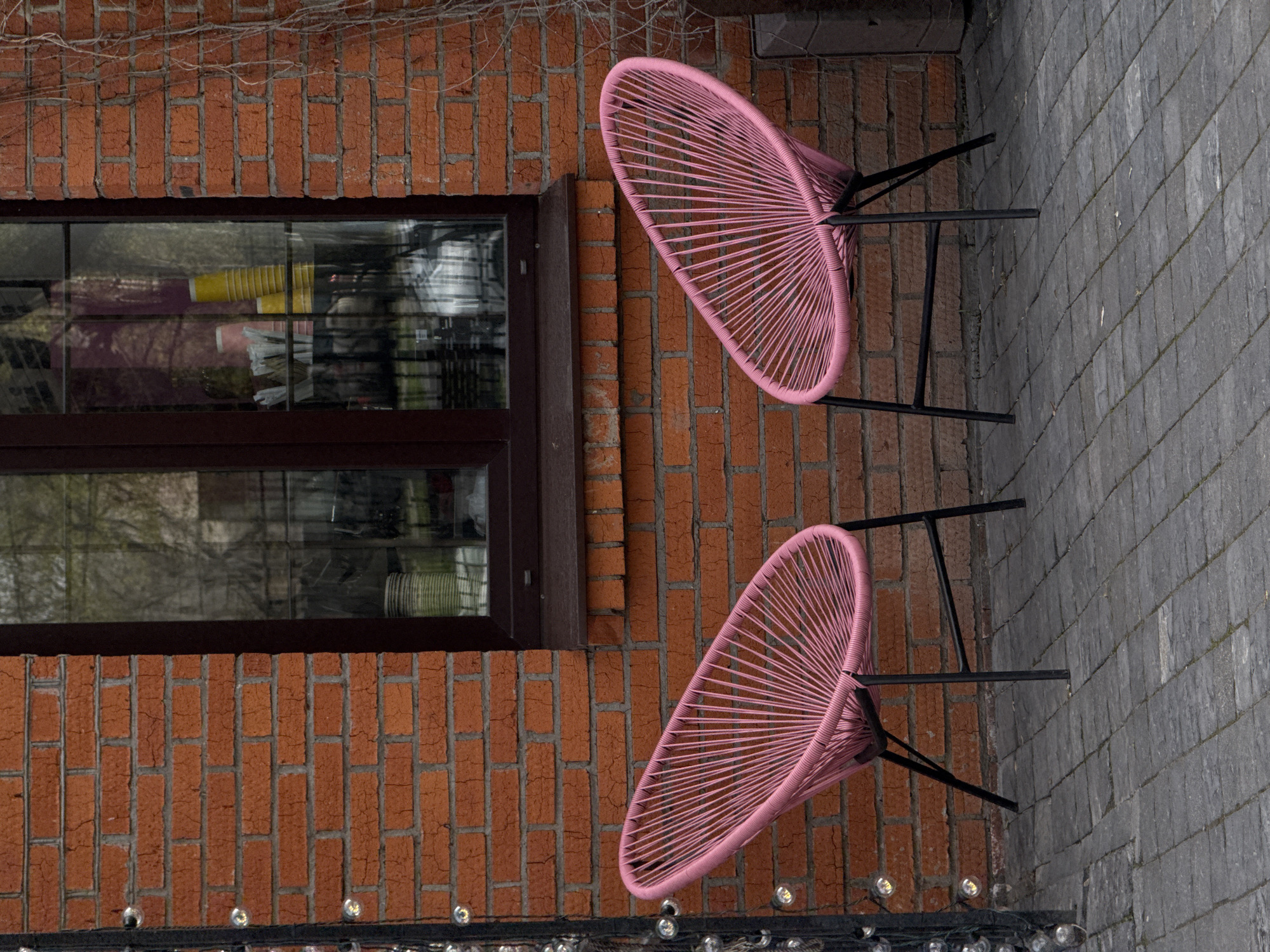 An outdoor scene captures a quiet patio or courtyard beside a brick building. Two vibrant pink Acapulco-style chairs, each featuring a woven plastic seat and a black metal frame, are positioned on a paved surface, seemingly ready for use. The chairs face away from the building, implying a view beyond the frame.
The building wall is constructed from reddish-brown bricks with contrasting grey mortar. A dark-framed window with multiple panes is set into this brick wall. Reflections within the window reveal glimpses of interior objects, including stacked yellow cups and white, possibly folded or rolled, items. Above the window, sparse, bare vines cling to the brickwork.
The ground is covered with rectangular grey pavers, providing a solid, textured surface. In the foreground, a string of clear, round-bulb lights is visible, suggesting ambient lighting for the evening, although the lights do not appear to be illuminated.
The lighting is soft and diffused, indicating an overcast day or possibly a transition period like dusk or dawn. There are no people visible, and no specific activity is taking place, creating a tranquil and inviting atmosphere. No text is visible within the image.