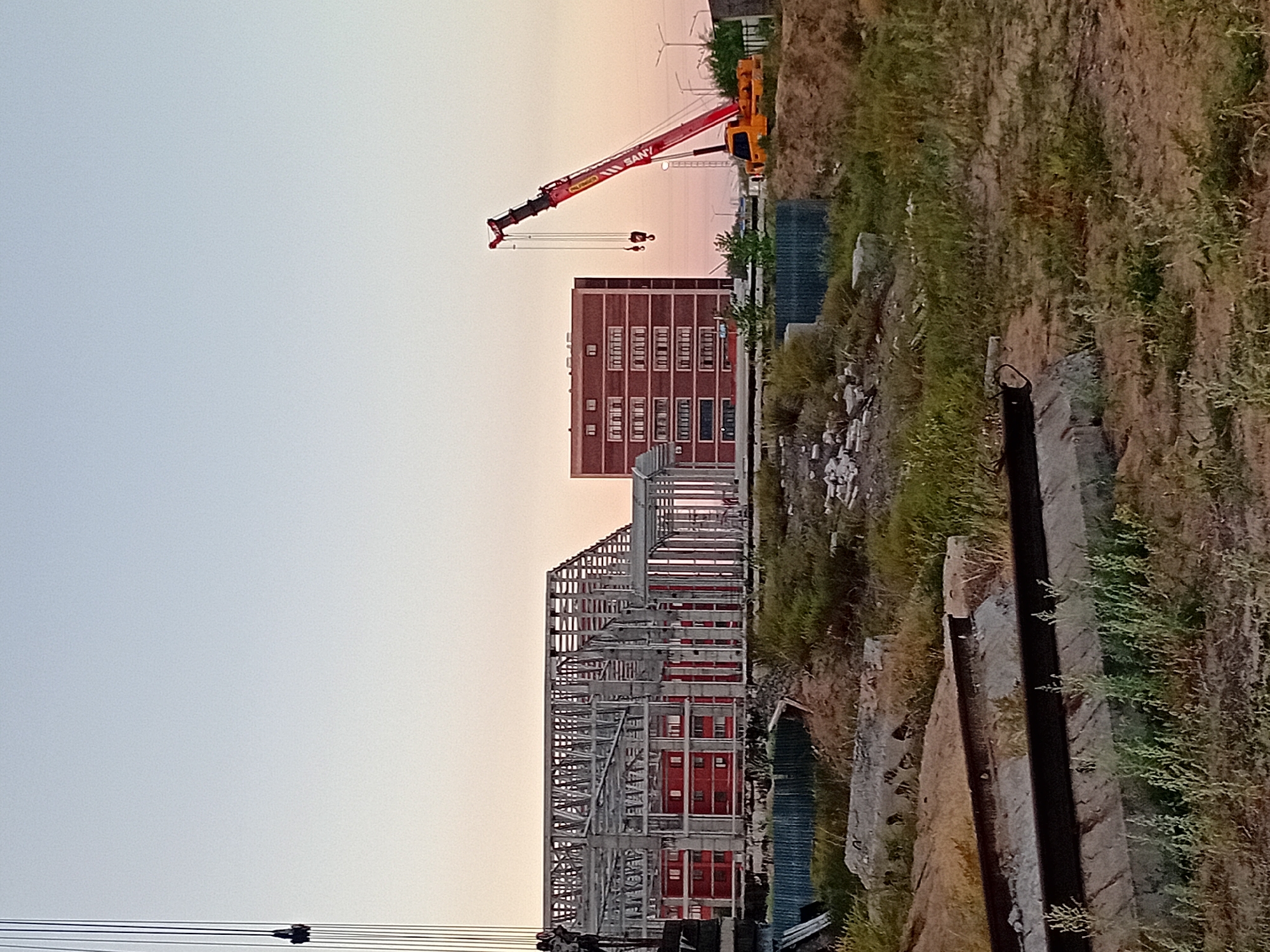The image depicts a construction site in the city of Semey, Kazakhstan, under a clear twilight sky. In the foreground, there are piles of concrete and steel beams, along with overgrown vegetation, suggesting an unfinished or abandoned area. Dominating the mid-ground is a large building under construction, characterized by its exposed metal framework and red brick walls. To the right, a bright red and yellow SANY brand crane is actively positioned, its boom extended towards the building, indicating ongoing work. Further in the background, a multi-story brick building with numerous windows stands complete, contrasting with the construction activity. The overall scene conveys a sense of progress and development in an urban environment.